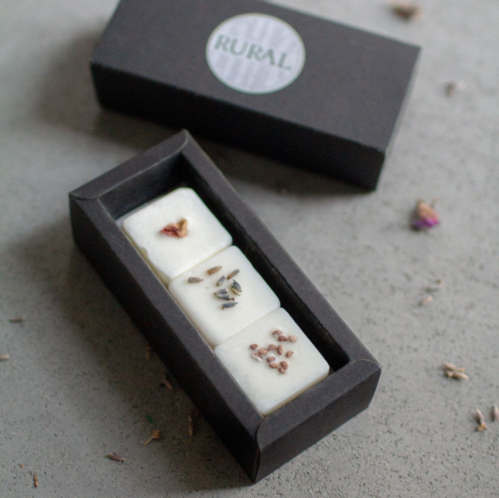 Close-up photo of a black gift box opened to show three square coconut soy wax melts handcrafted by RURAL Candles, decorated with red and purple flower petals and tiny blackberry seeds. All artfully displayed on a grey stone background.