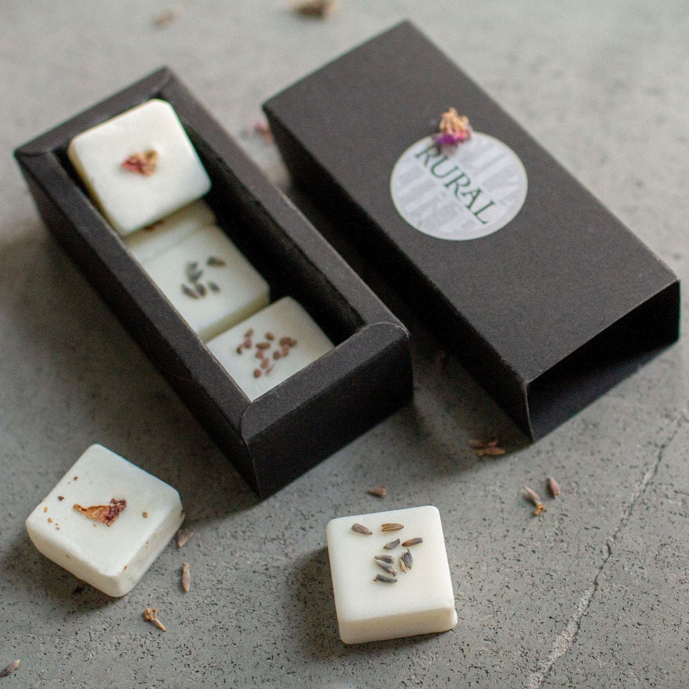 Photo of a black gift box opened to show six square coconut soy wax melts handcrafted by RURAL Candles, decorated with red and purple flower petals and tiny blackberry seeds. All artfully displayed on a grey stone background.