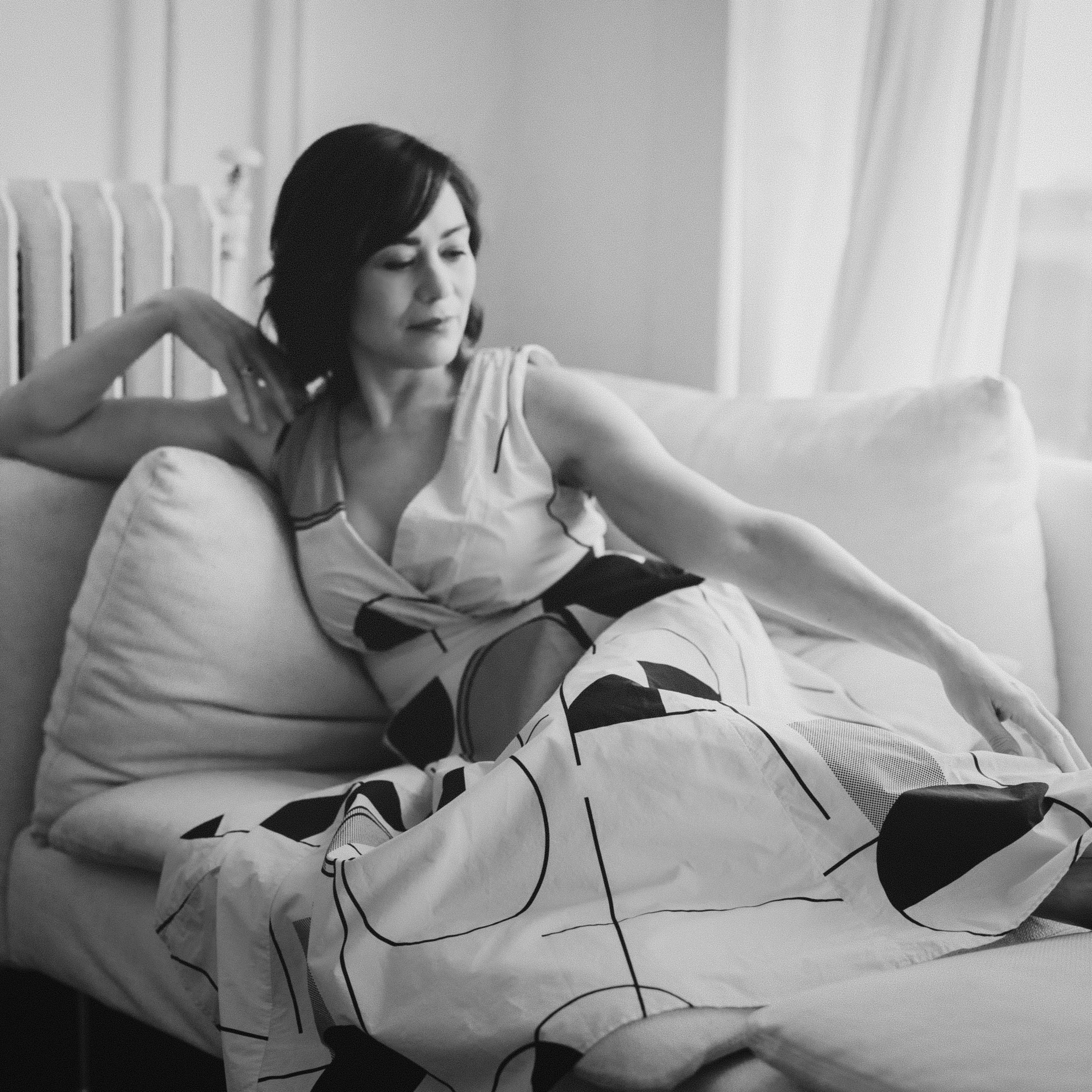 Black and white photo of Monique, the owner and chief candlemaker at RURAL Candles. She's reclining on a white sofa, barefoot, in a romantic, long dress with an art deco pattern on it. Vibe is very vintage beauty.