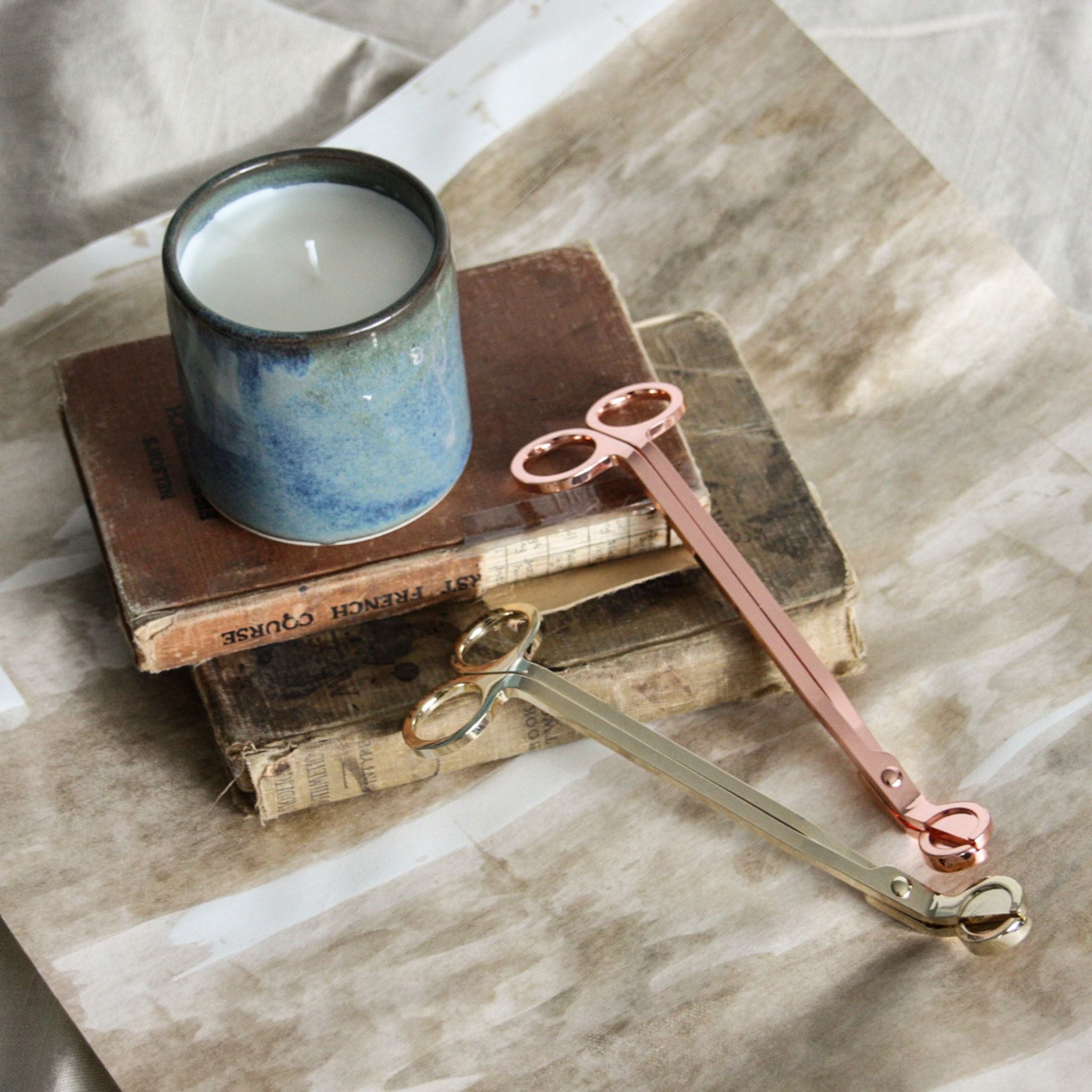Photo of a beautiful blue handmade pottery candle, sitting on top of vintage books and art paper. Two sets of candle wick trimmers are displayed alongside the candle, one is rose gold and the other is gold. Looks like the photo is in a cool art studio.