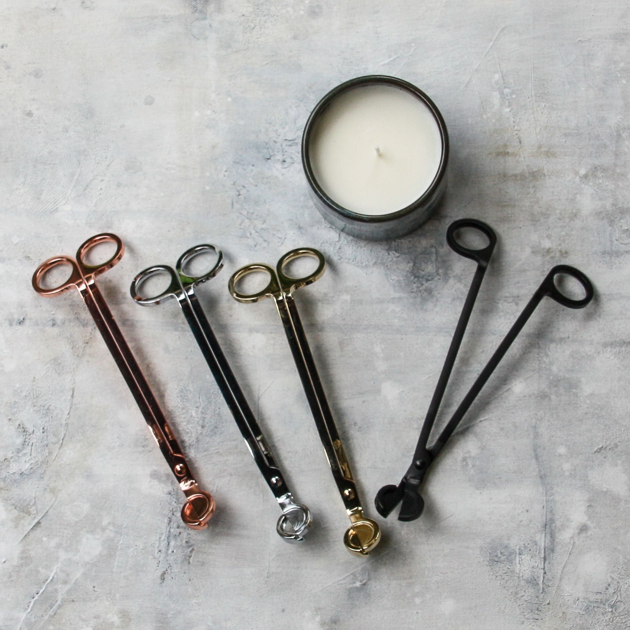 Photo of four candle wick trimmers in rose gold, silver, gold and black, alongside a handcrafted pottery candle, on top of a grey paint canvas. Vibe is very art studio cool.