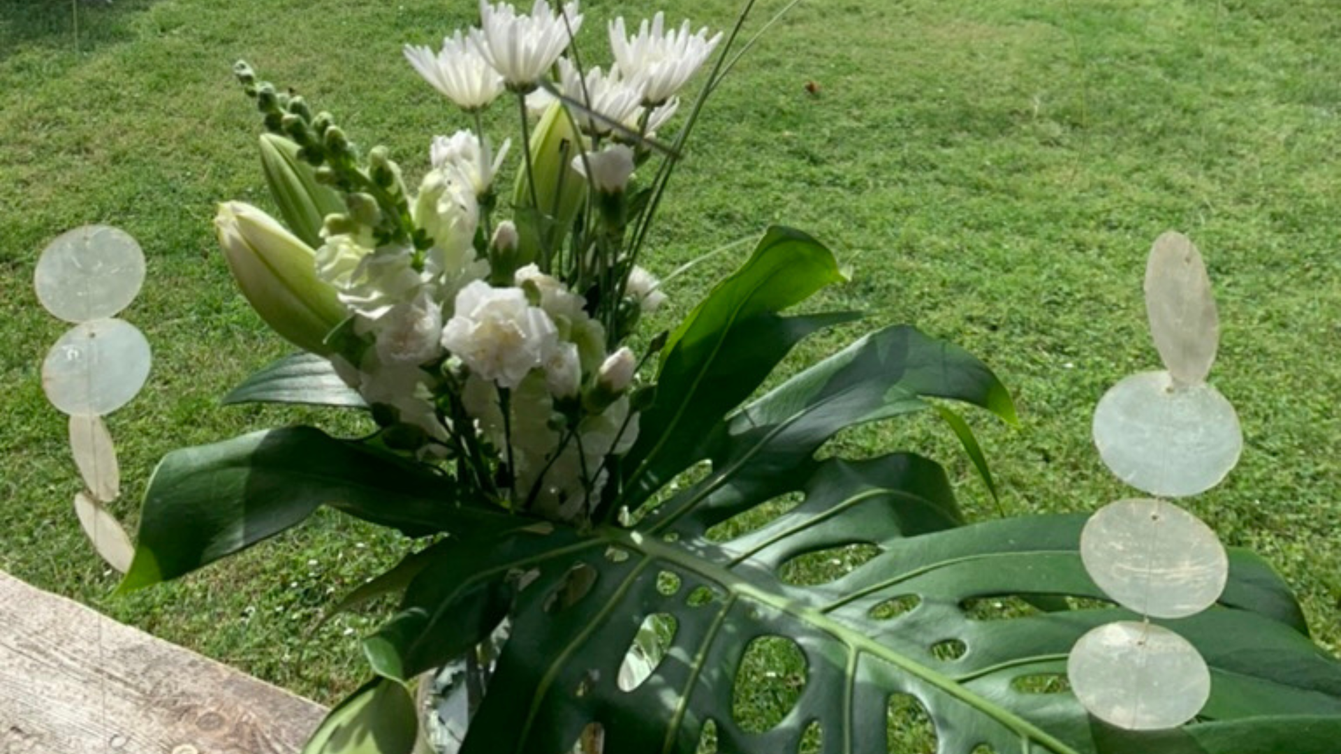 Photo of a white floral bouquet with large exotic leaves and seashells, on a green grass background.