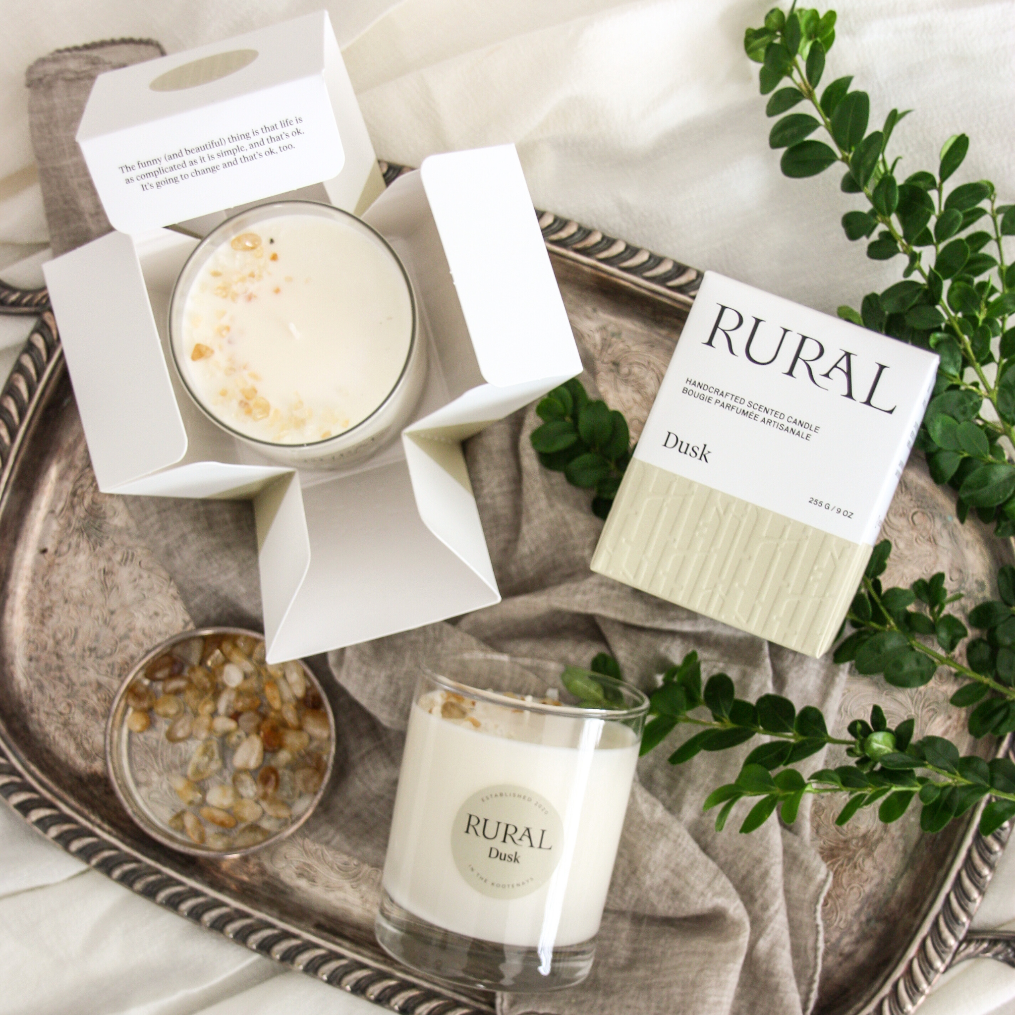 Top view photo of two white candles in clear glass with round tan labels indicating RURAL Candles Dusk Scent beside its matching tan and white elegantly embossed candle gift box. We can see the top of the candle is decorated with small gold colored gemstones, artfully arranged in a crescent moon shape.