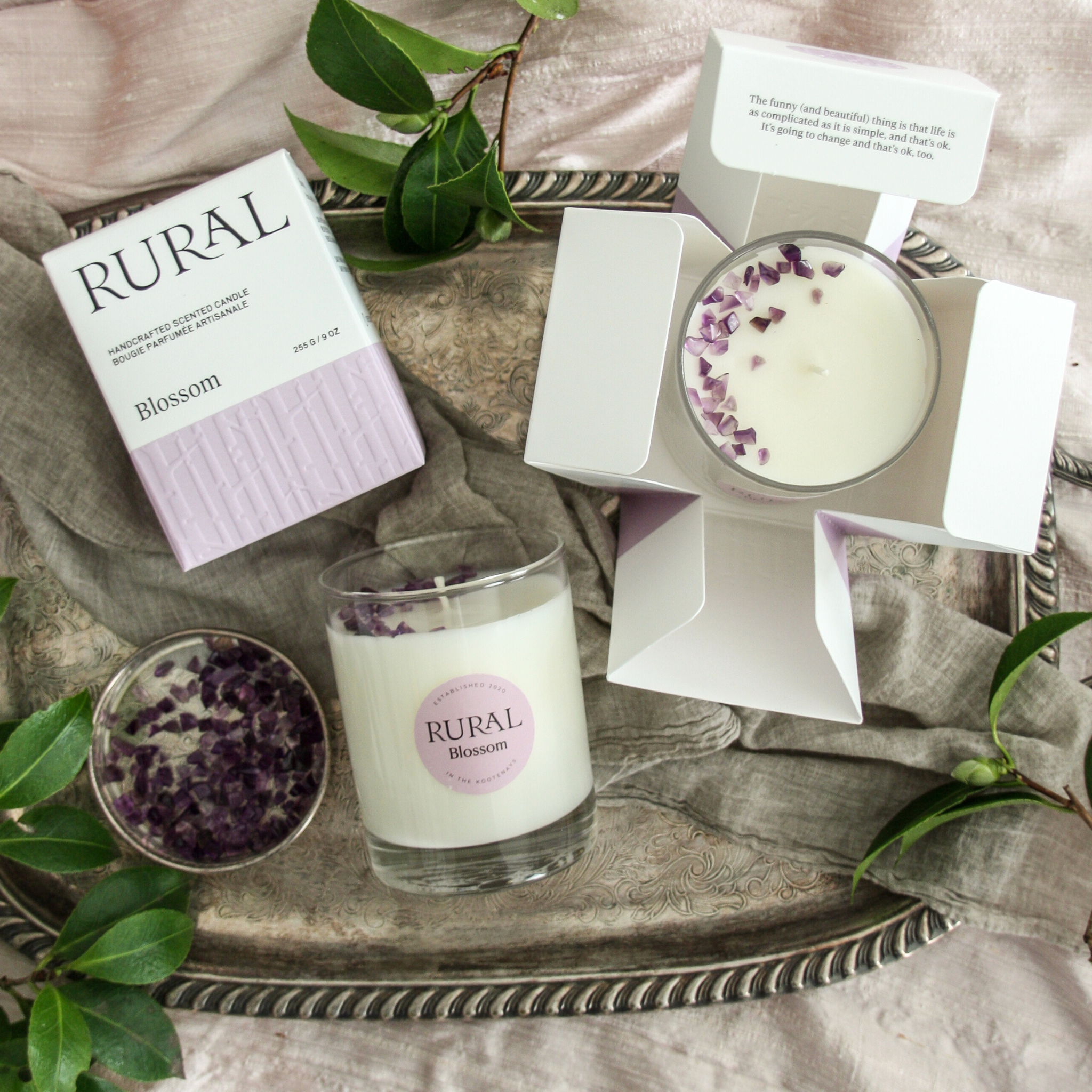Photo of a white candle in a clear glass container with a light purple round RURAL Candles label on it, indicating the scent is called Blossom. The candle top is embellished with a sprinkling of purple amethyst chips, and the candle is sitting on a silver tray with grey linen, pink silk and green foliage in the background. We can also see the candle in its matching purple and white gift box, which opens like a flower.