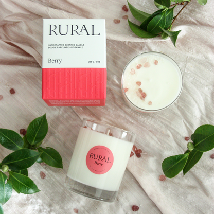 Photo of two white candles in clear glass containers with a pink round RURAL Candles label on, indicating its Berry scent. The candles are on a pink silk fabric with green foliage decorating the background. The tops of the candles are sprinkled with various shades of pink strawberry quartz crystals. Alongside is a matching pink and white Berry Candle gift box with the RURAL Candles logo on it.