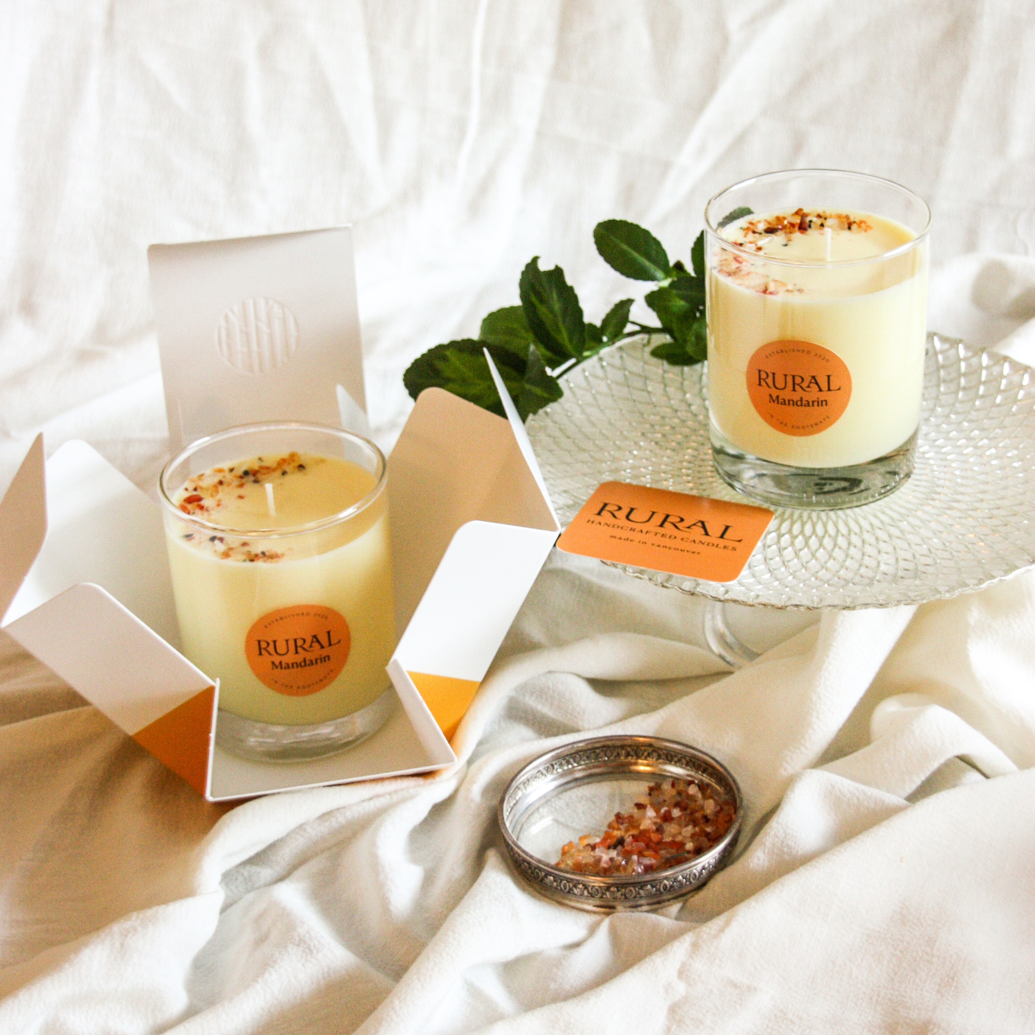 Photo of two candles in clear glass with a round orange label indicating RURAL Candles Mandarin orange scent beside its matching orange and white elegantly embossed candle gift box. We can see the top of the candles are decorated with small orange carnelian gemstones, artfully arranged in a crescent moon shape, and the gift box opens like a flower.