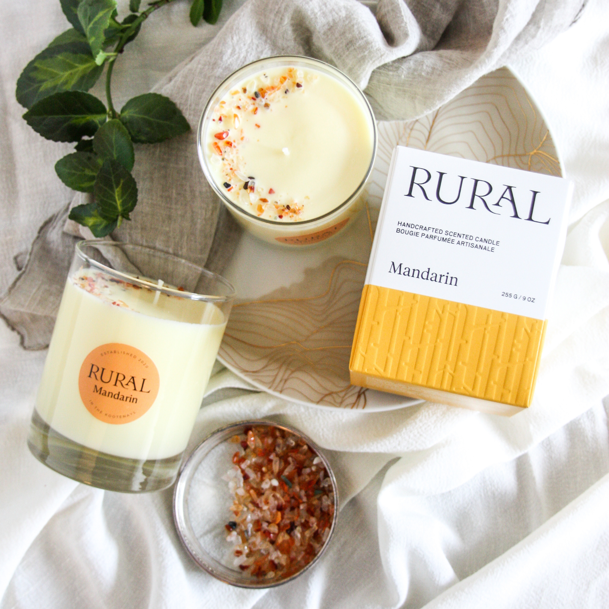 Top view photo of two candles in clear glass with a round orange labels indicating RURAL Candles Mandarin Scent beside its matching orange and white elegantly embossed candle gift box. We can see the top of the candle is decorated with small orange carnelian gemstones, artfully arranged in a crescent moon shape. The setting is very elegant and inviting Old World kitchen style.