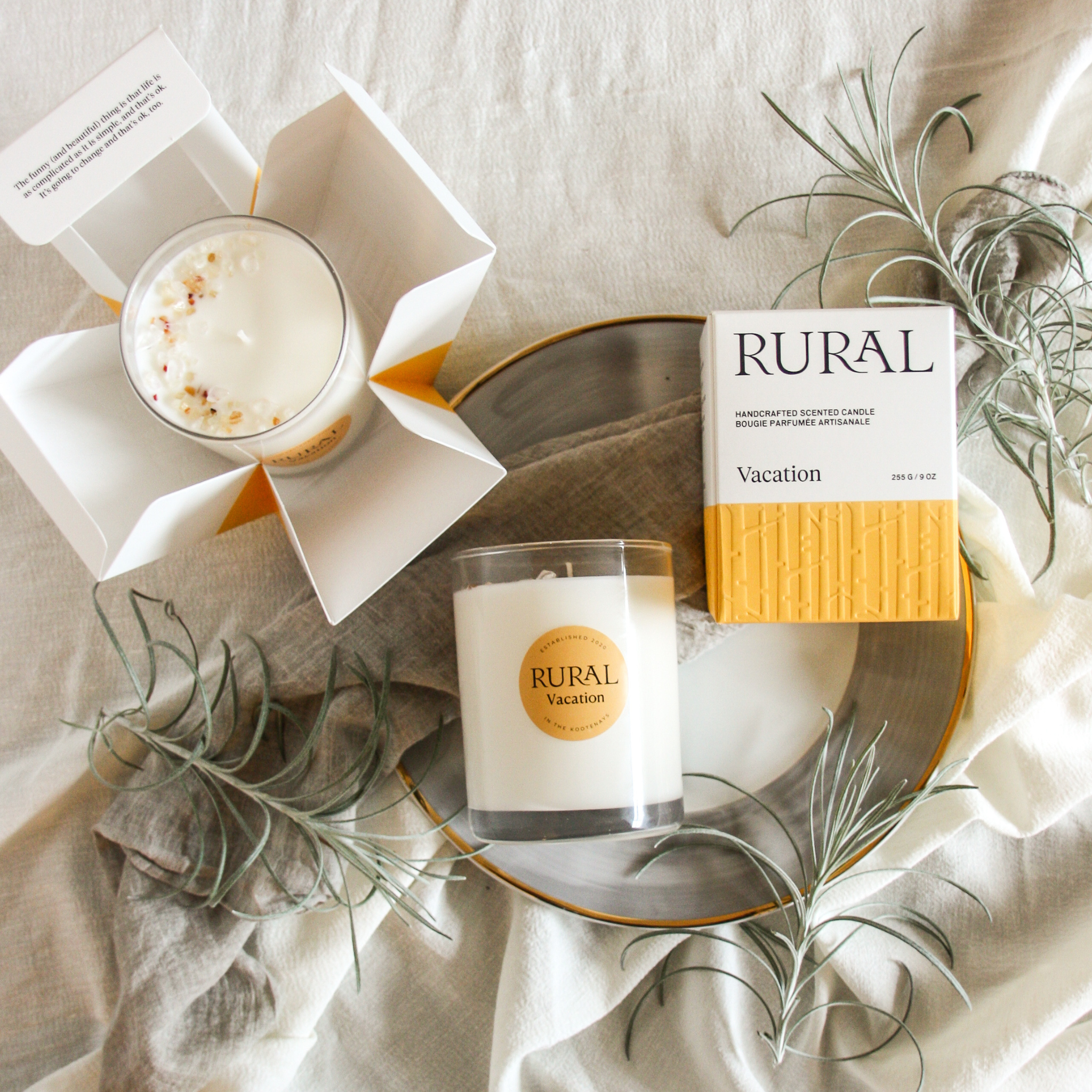 Photo of two white candles in clear glass containers with the round RURAL Candles label on it, indicating the Vacation scent. We can see the tops of the candles are sprinkled with orange carnelian gemstone chips in a crescent moon shape, and the matching orange and white RURAL Candles gift box is beautifully embossed. The candles are sitting artfully displayed on a grey platter with grey and ivory linens underneath and green herbal sprigs scattered around. The scene is a very calm and earthy.