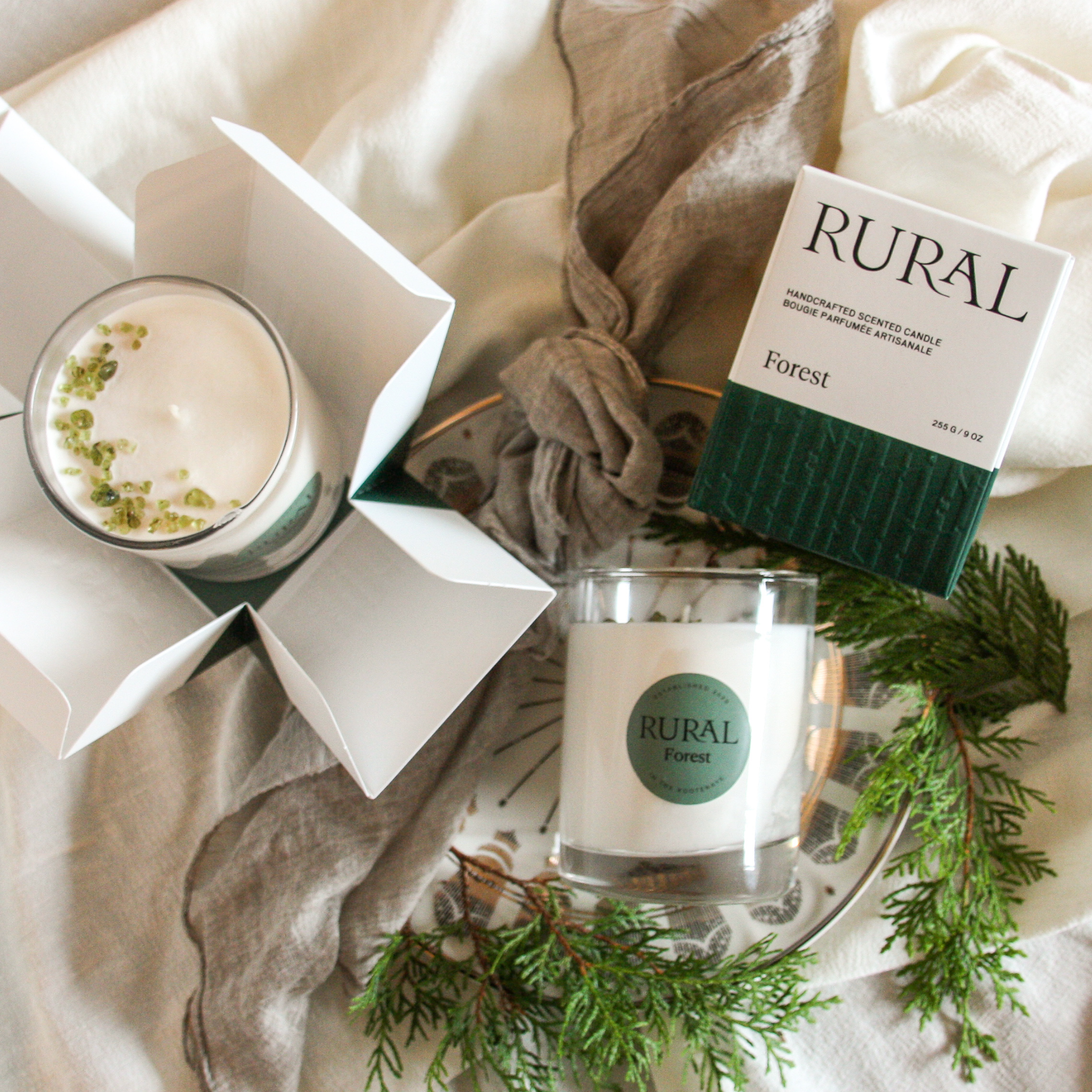 Top view photo of two white candles in clear glass with a round green label indicating RURAL Candles Forest Scent beside its matching green and white elegantly embossed candle gift box. We can see the top of the candle is decorated with small green gemstones, artfully arranged in a crescent moon shape. The candles are arranged on earthy toned linens with cedar branches alongside. 