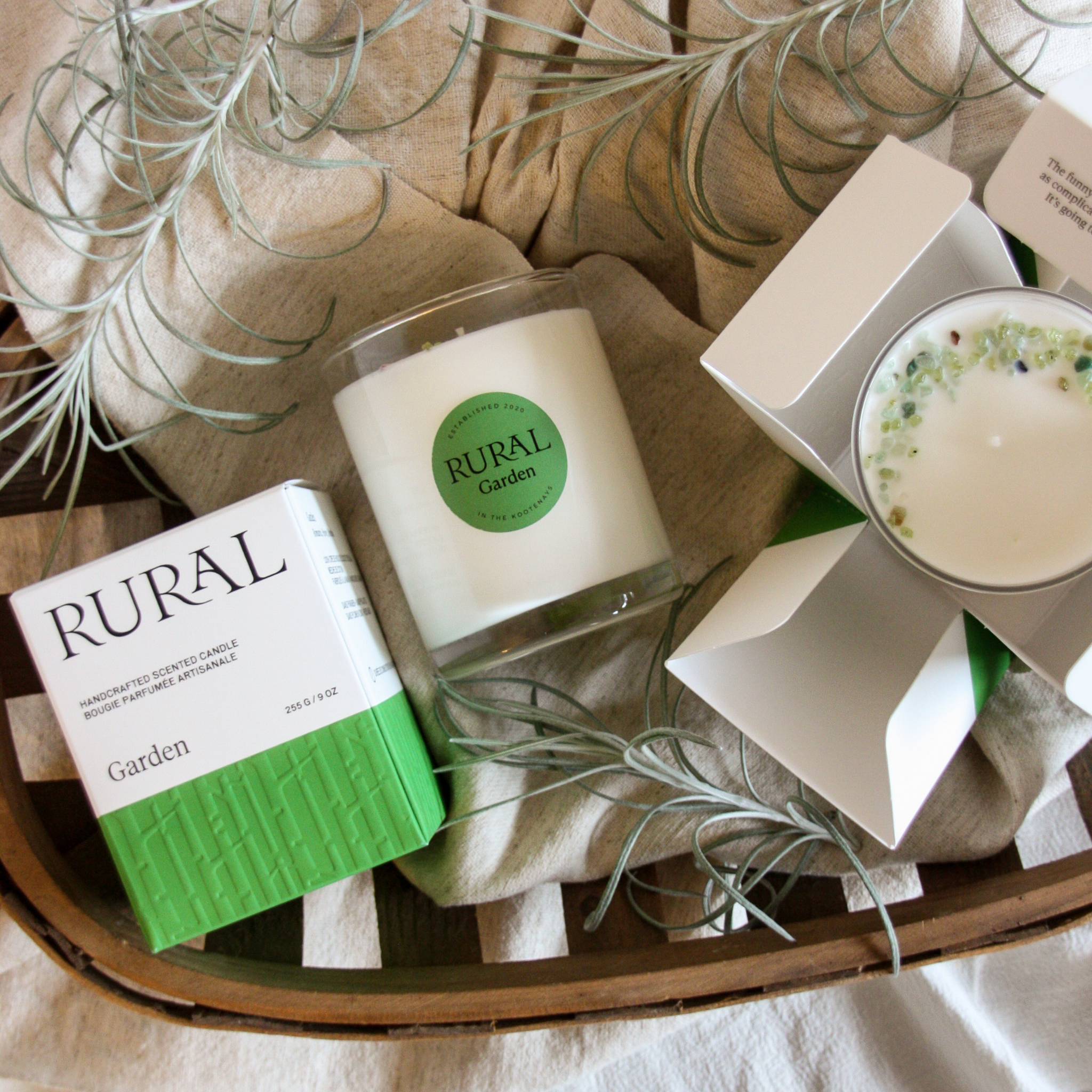 Photo of two coconut soy candles in clear white glasses with a round green label indicating RURAL Candles Garden Scent. The gemstone embellished candles are sitting on a wooden tray with grey linen and wispy green grasses surrounding it. Alongside is a matching green and white RURAL Candle gift box, which is embossed and elegant.