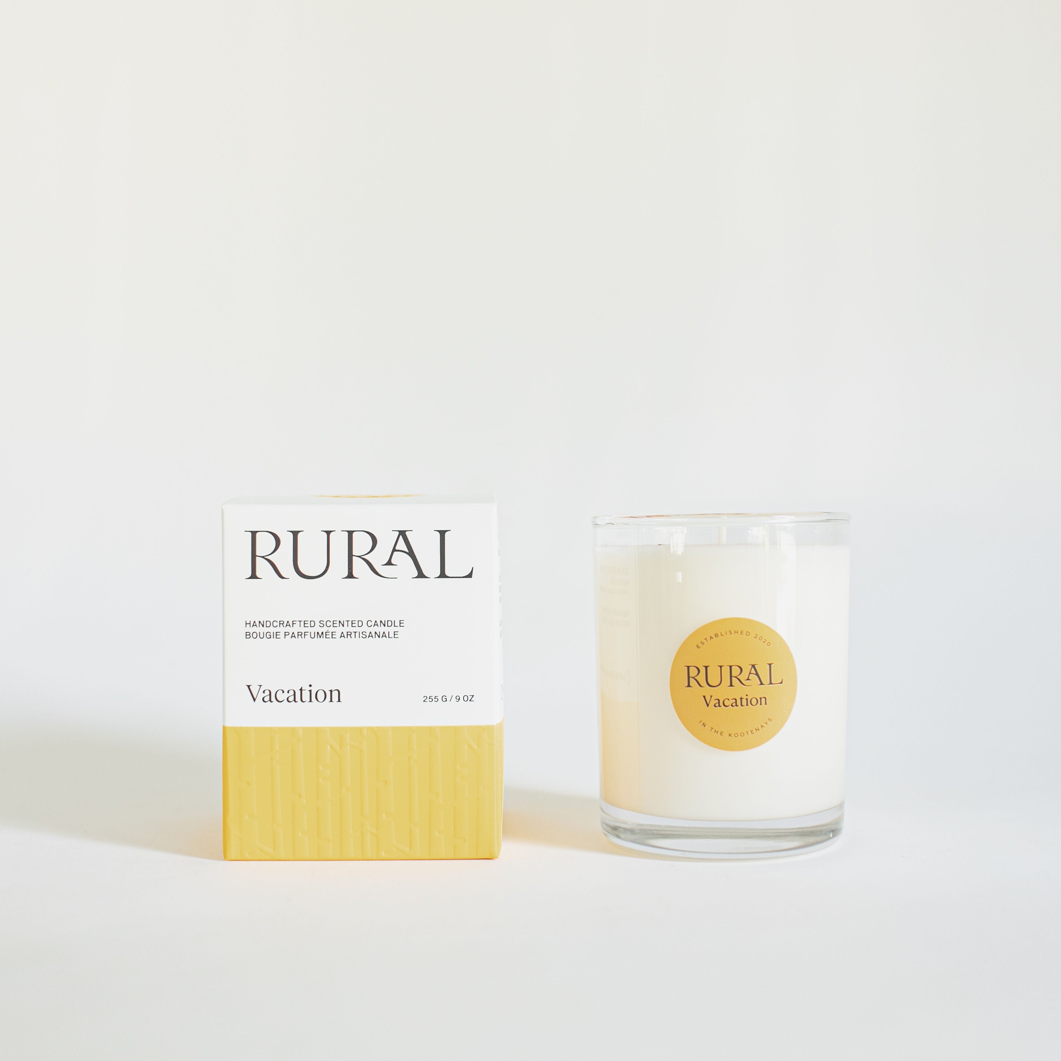 Product Photo of a white candle in a clear glass container with the round RURAL Candles label on it, indicating the Vacation scent alongside its matching orange and white RURAL Candles gift box, which is beautifully embossed. 