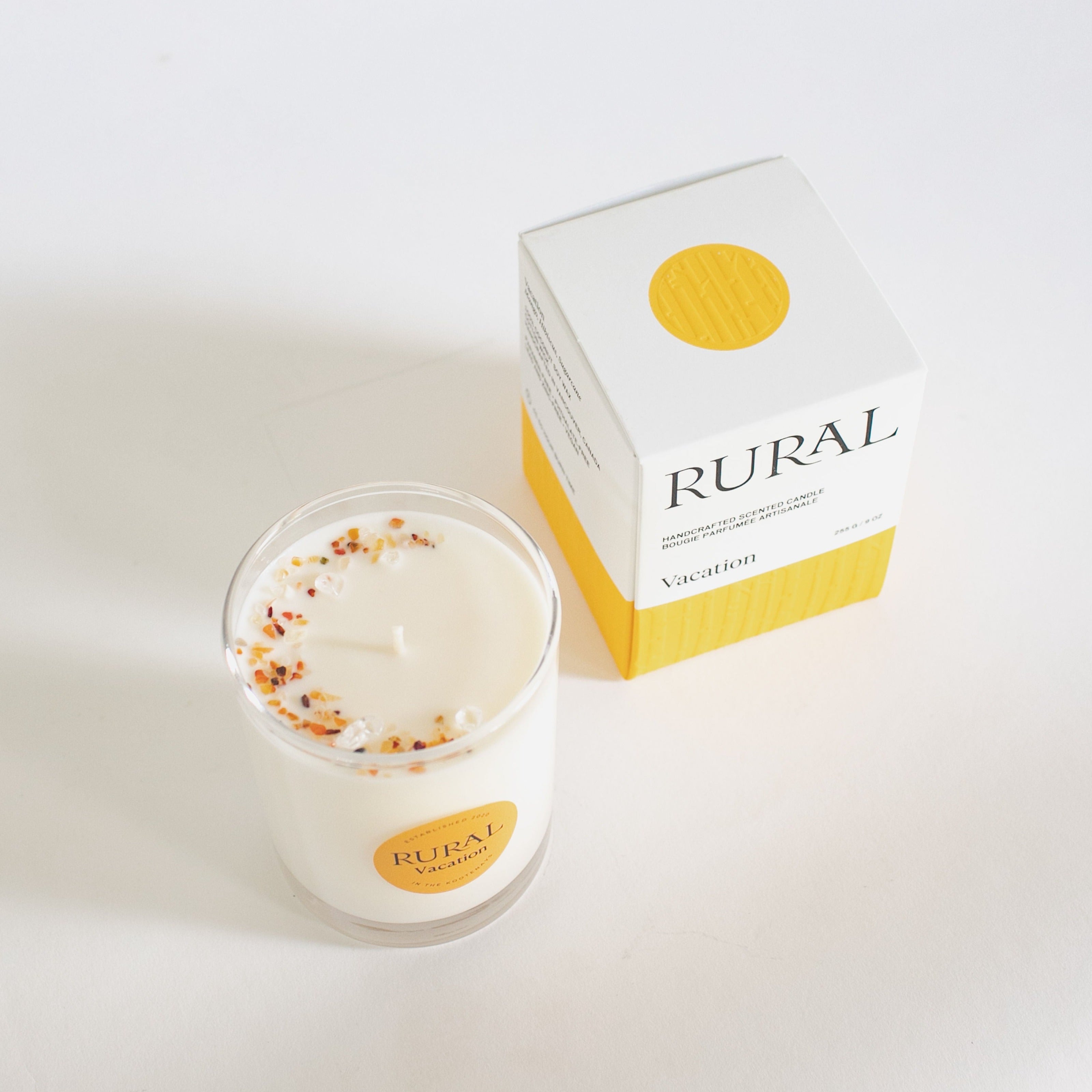 Top view product photo of a white candle in a clear glass container with the round RURAL Candles label on it, indicating the Vacation scent. We can see the top of the candle is sprinkled with orange carnelian gemstone chips in a crescent moon shape, and the matching orange and white RURAL Candles gift box is beautifully embossed. 