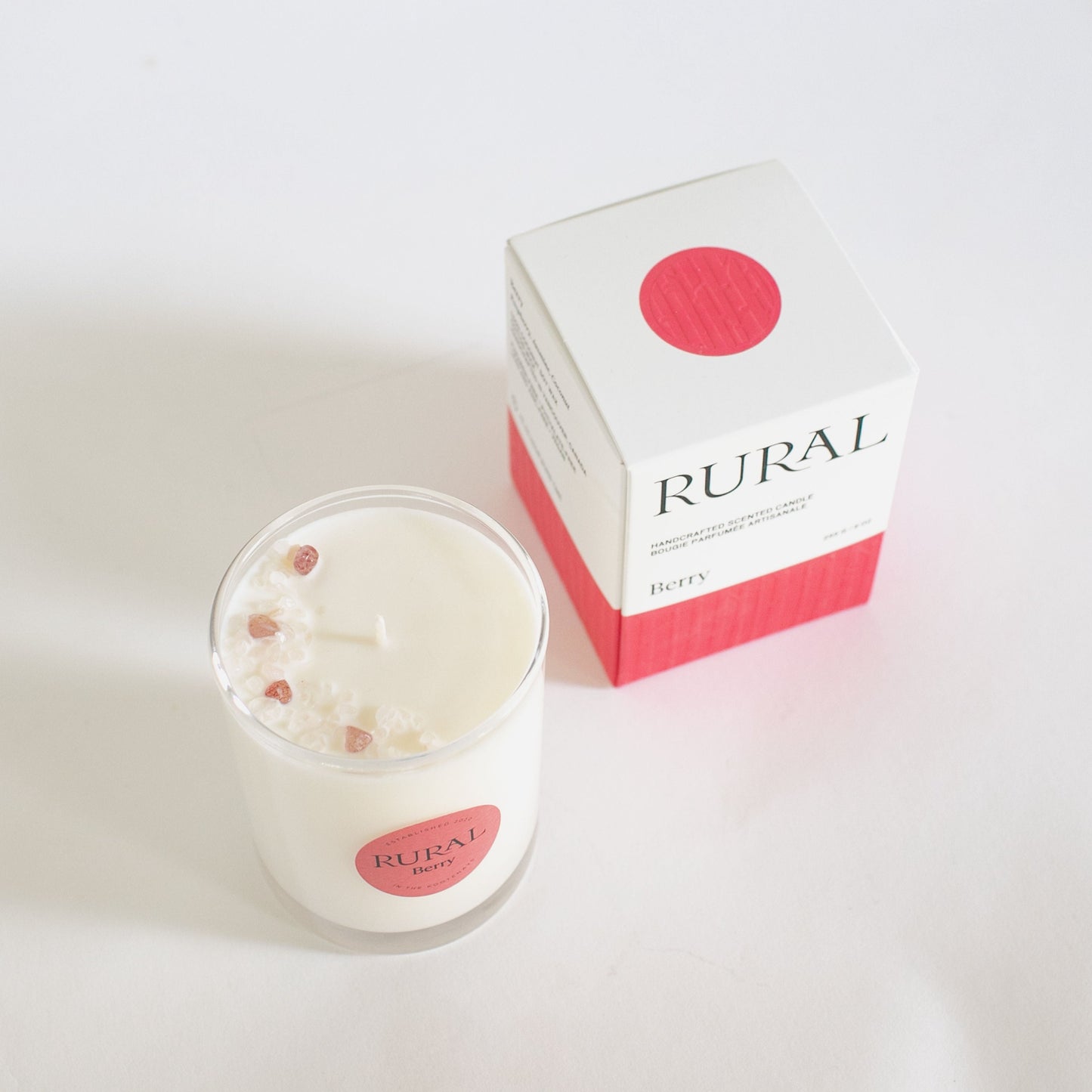 Top view product photo of a white candle in clear glass container with a pink round RURAL Candles label on, indicating its Berry scent. The top of the candle is decorated with small pink and white strawberry quartz crystals, arranged in the shape of a crescent moon. Alongside is a matching pink and white embossed Berry Candle gift box with the RURAL Candles logo on it.