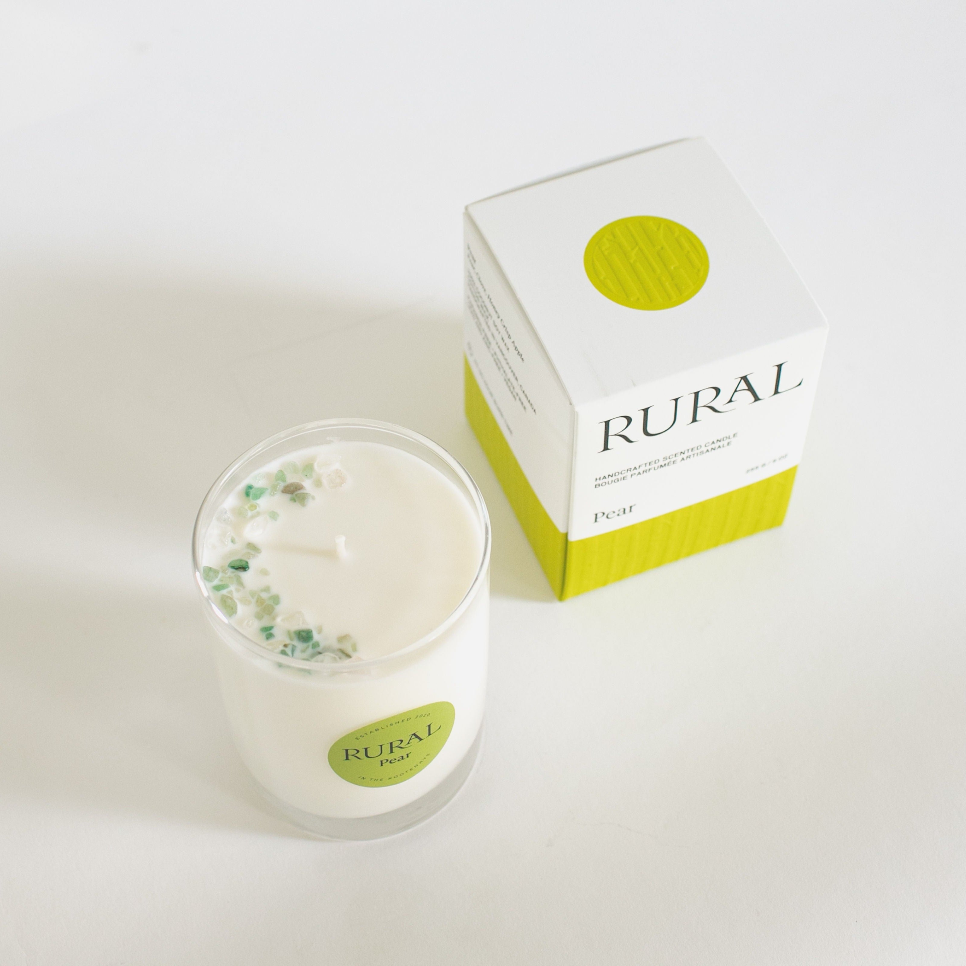 Top view photo of a white candle in a clear glass with a round green label indicating RURAL Candles Pear Scent beside its matching green and white elegantly embossed candle gift box. We can see the top of the candle is decorated with small green gemstones, artfully arranged in a crescent moon shape.
