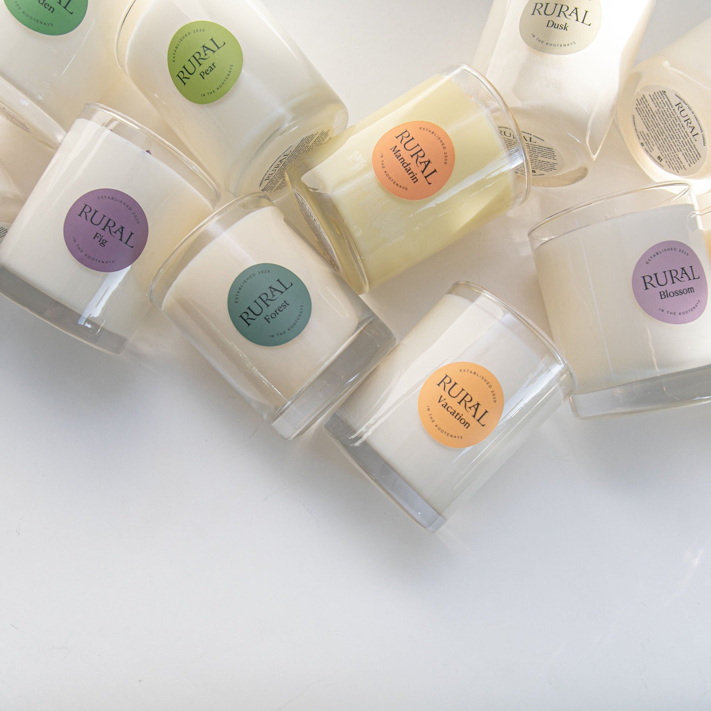 Photo of a group of white candles in clear glass with multicolor labels. RURAL Candles in scents such as Fig, Pear, Mandarin, Blossom, Forest, Dusk, Garden and Vacation. The vibe is clean and calm yet playful.