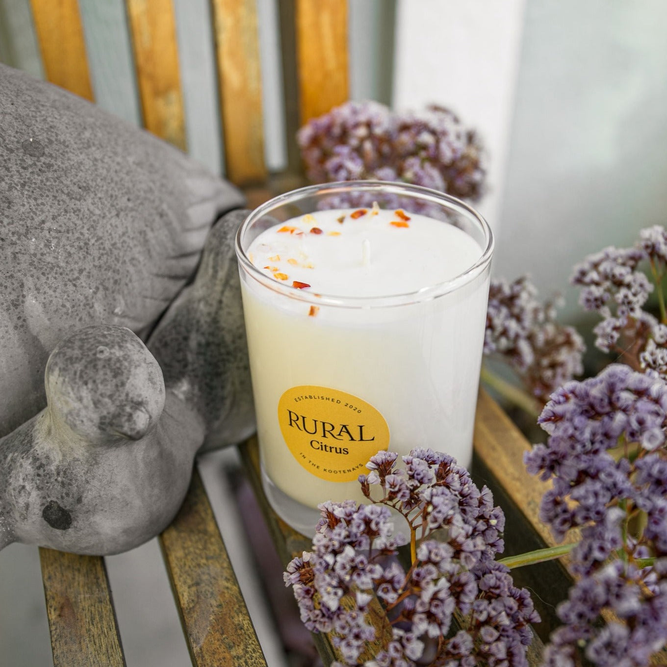 Top view photo of a white candle in a clear glass container with a yellow round RURAL Candles label on it, indicating the scent is called Citrus. The Citrus candle looks fresh and inviting, sitting on a wooden outdoor chair alongside a grey statue of a little bird, and some purple flowers.