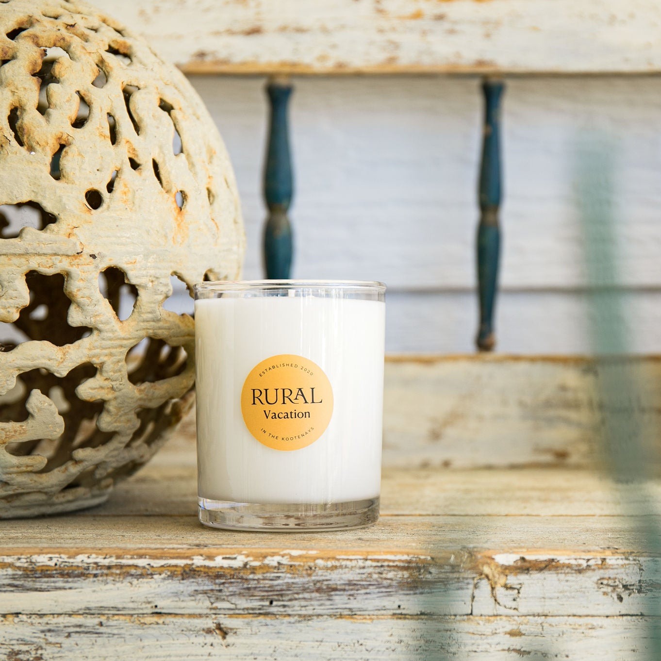 Photo of a white candle in a clear glass container with the round RURAL Candles label on it, indicating the Vacation scent. The candle is sitting on a rustic wooden bench beside a round metal garden ornament, and the scene is a very serene and inviting.