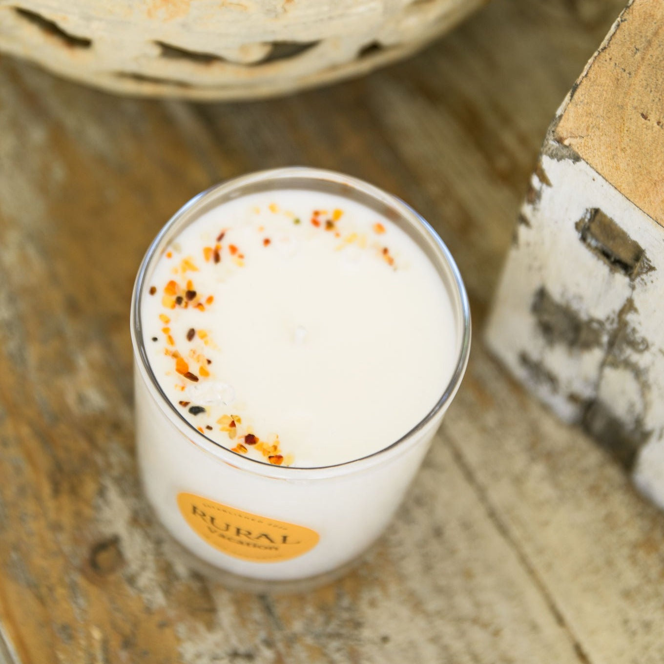 Top view photo of a white candle in a clear glass container with the round RURAL Candles label on it, indicating the Vacation scent. The candle is sitting on a rustic wooden bench beside some matching garden ornaments, and we can see the top of the candle is sprinkled with orange carnelian gemstone chips in a crescent moon shape. The scene is a very serene and inviting.
