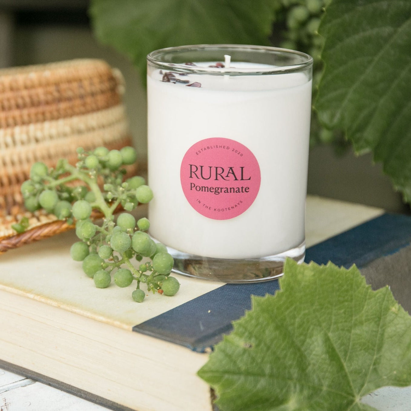 Photo of a white candle in a clear glass with a round red label indicating RURAL Candles Pomegranate Scent. The candle is on a book beside some green grapes, wicker and lush leaves. The scene is a fresh garden on a summer's day.