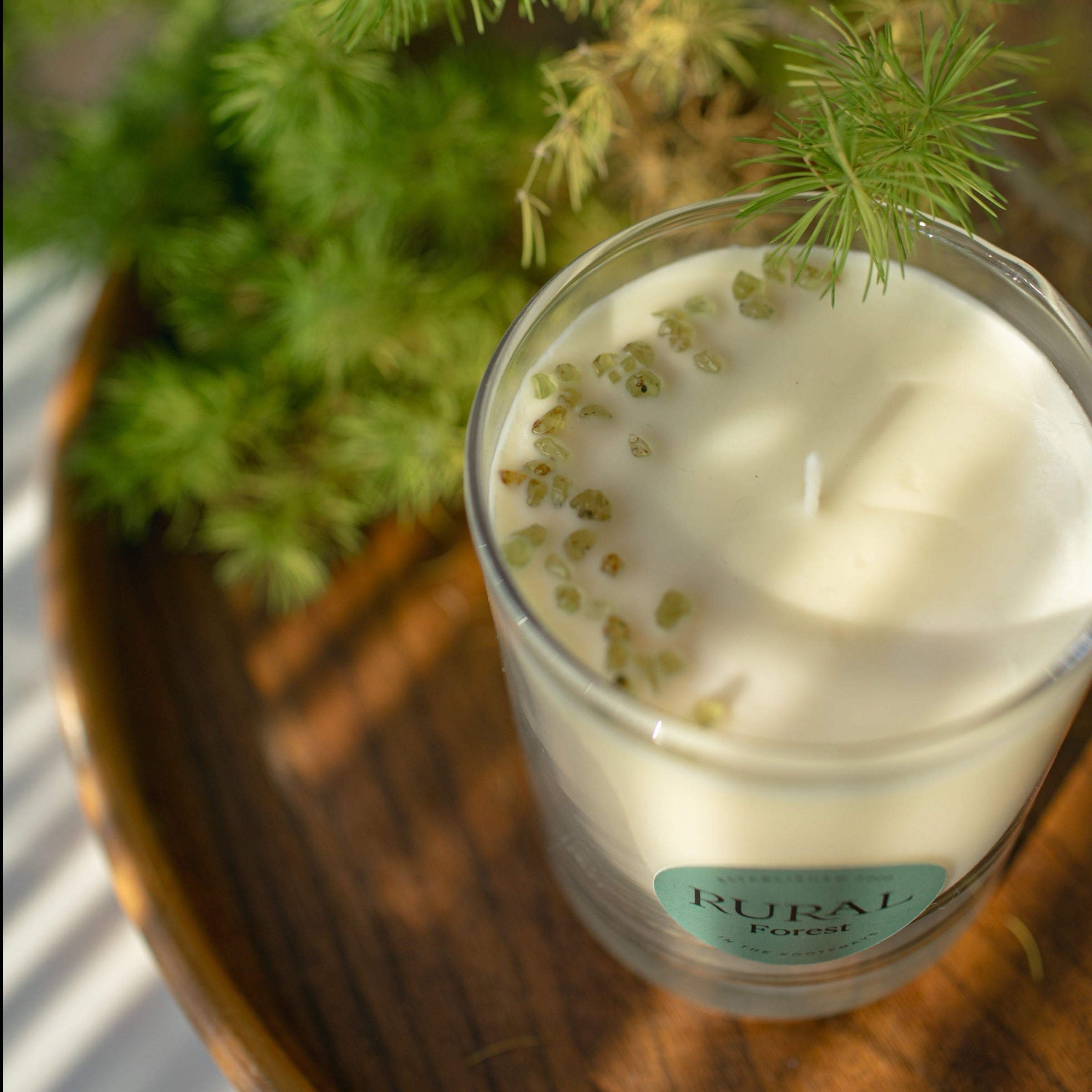 Top view photo of a white candle in a clear glass with a round green label indicating RURAL Candles Forest Scent. The candle is on a wooden tray with green foliage surrounding, suggesting the outdoors, perhaps a walk in the woods with pine and fir trees. We can see the top of the candle is embellished with small green olivine gemstones arranged in a crescent moon shape.