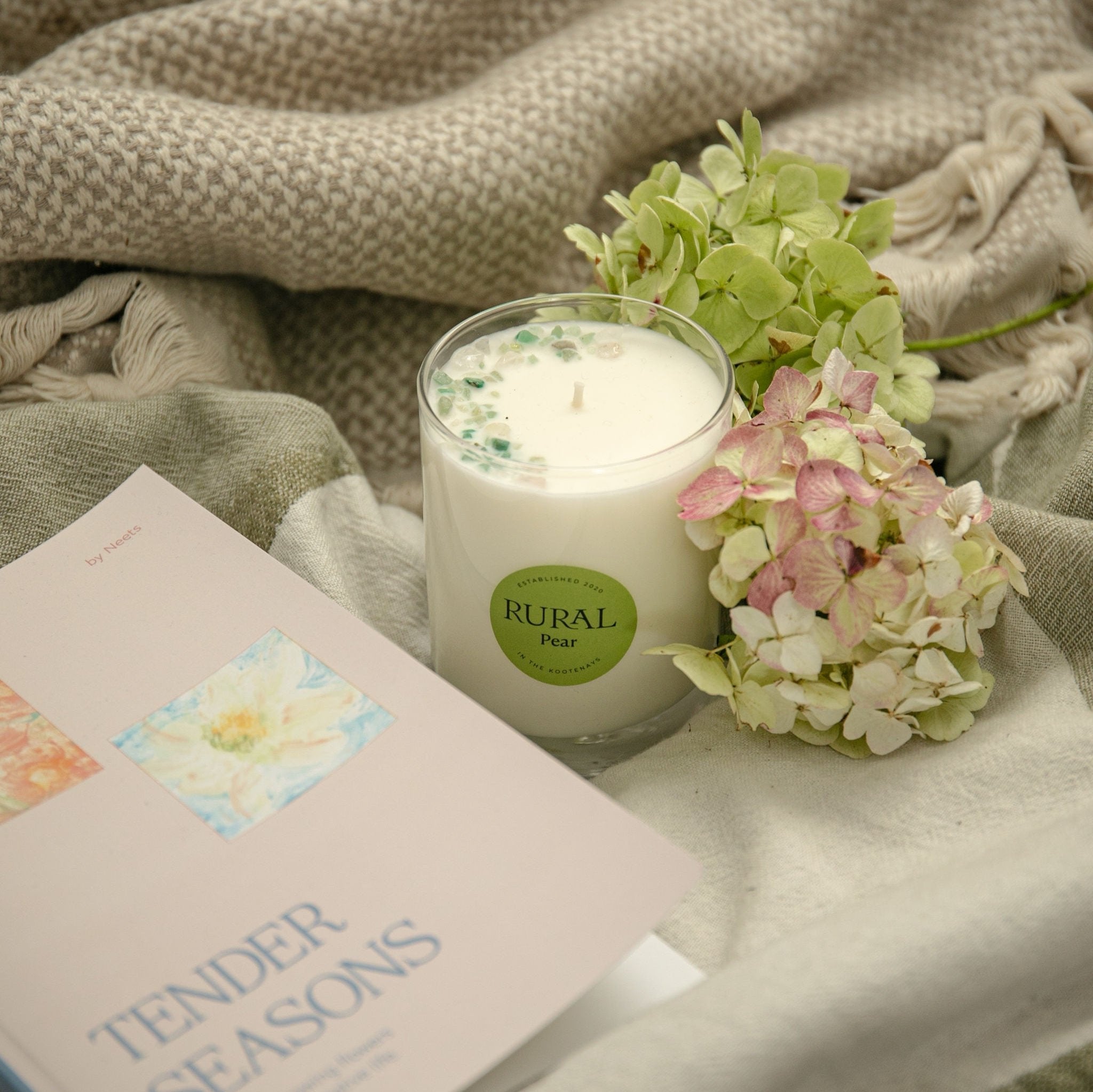Photo of a white candle in a clear glass with a round green label indicating RURAL Candles Pear Scent. We can see the top of the candle is decorated with small green gemstones, artfully arranged in a crescent moon shape. The candle is surrounded by a book, titled Tender Seasons, cozy throws, and flowers, very comforting and inviting outdoor space.