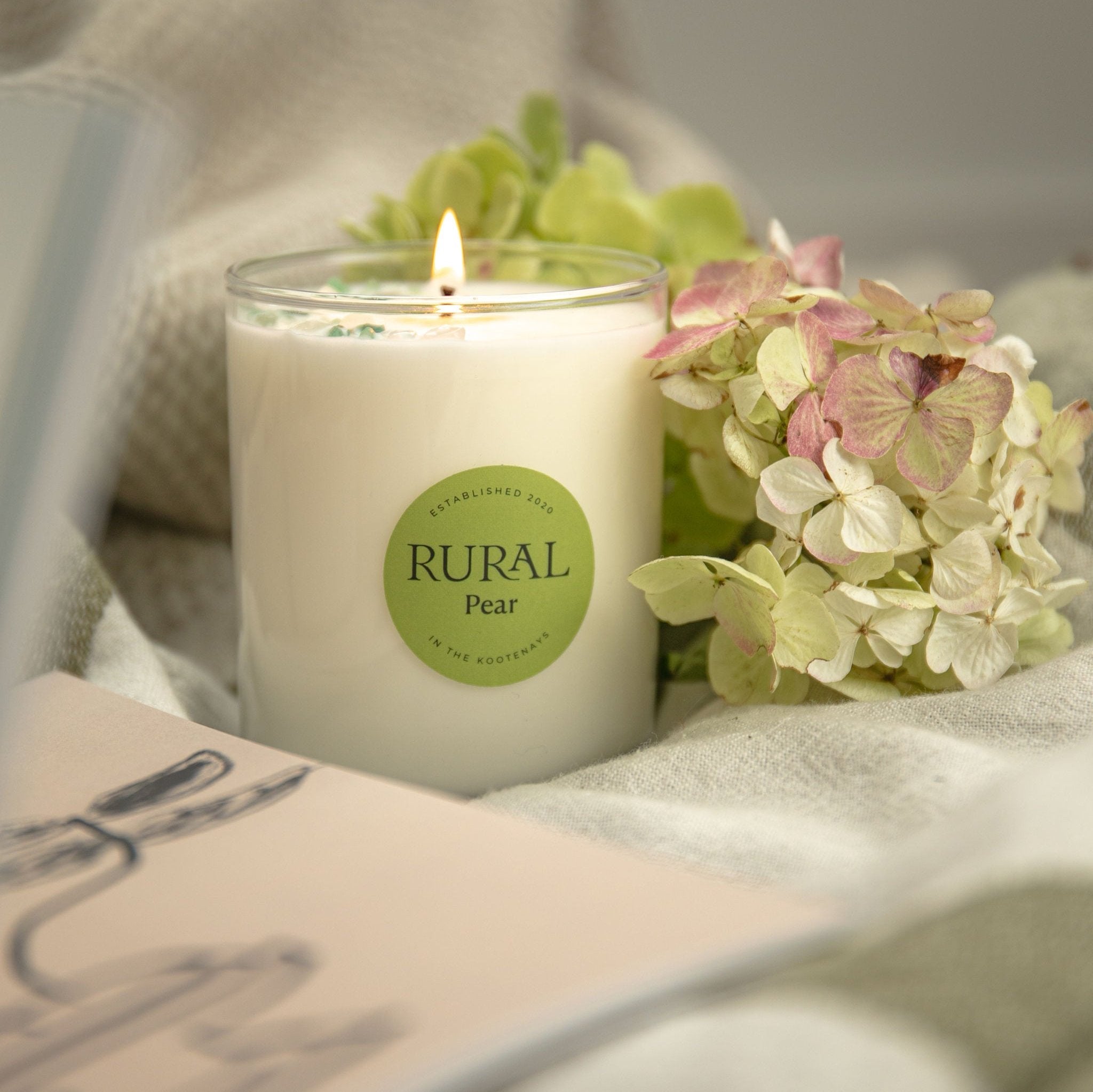 Photo of a lit white candle in a clear glass with a round green label indicating RURAL Candles Pear Scent. The candle is surrounded by flowers and books on a linen cloth, very romantic and clean candle scene.