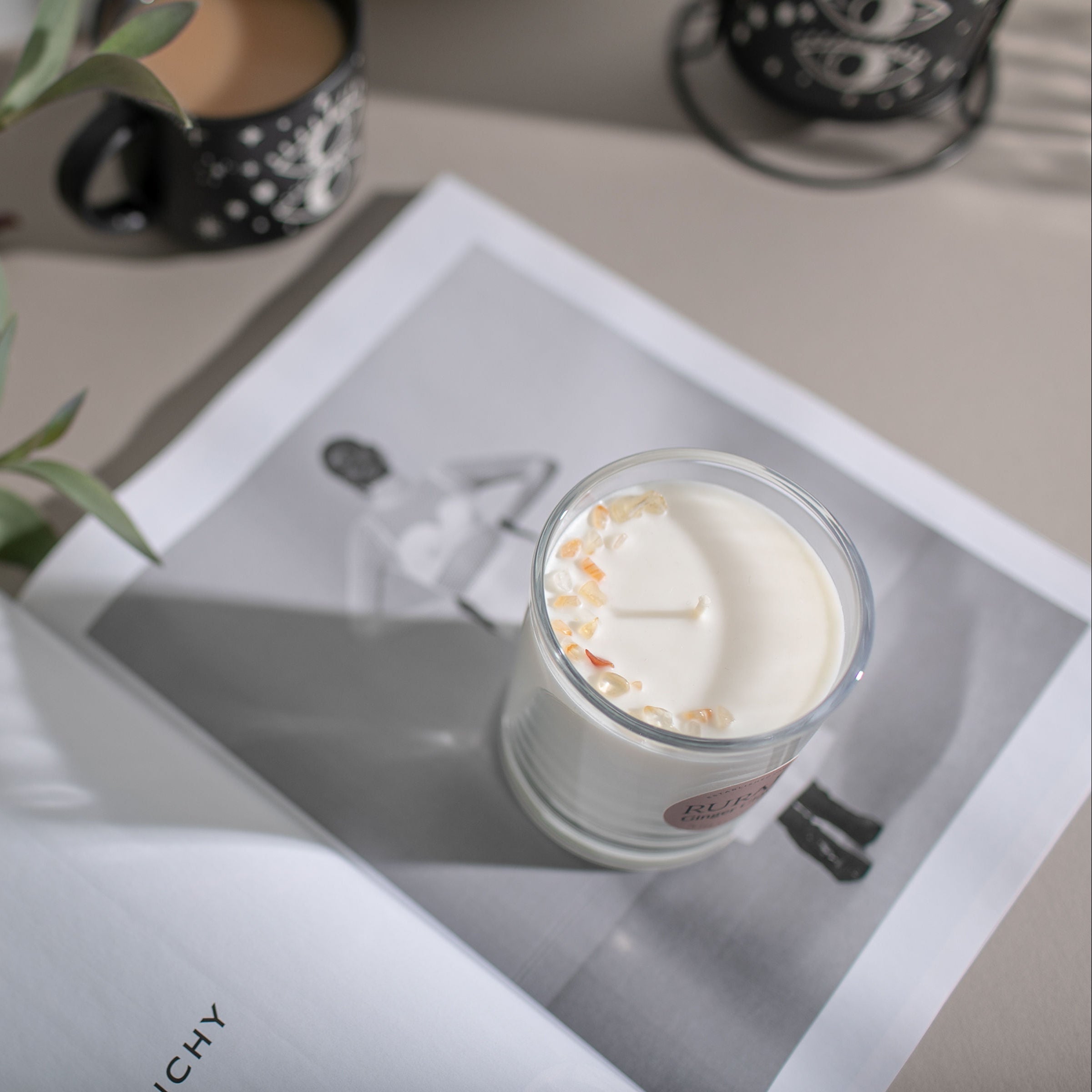 A candle in a glass jar on a white book surrounded by a plant and a cup.