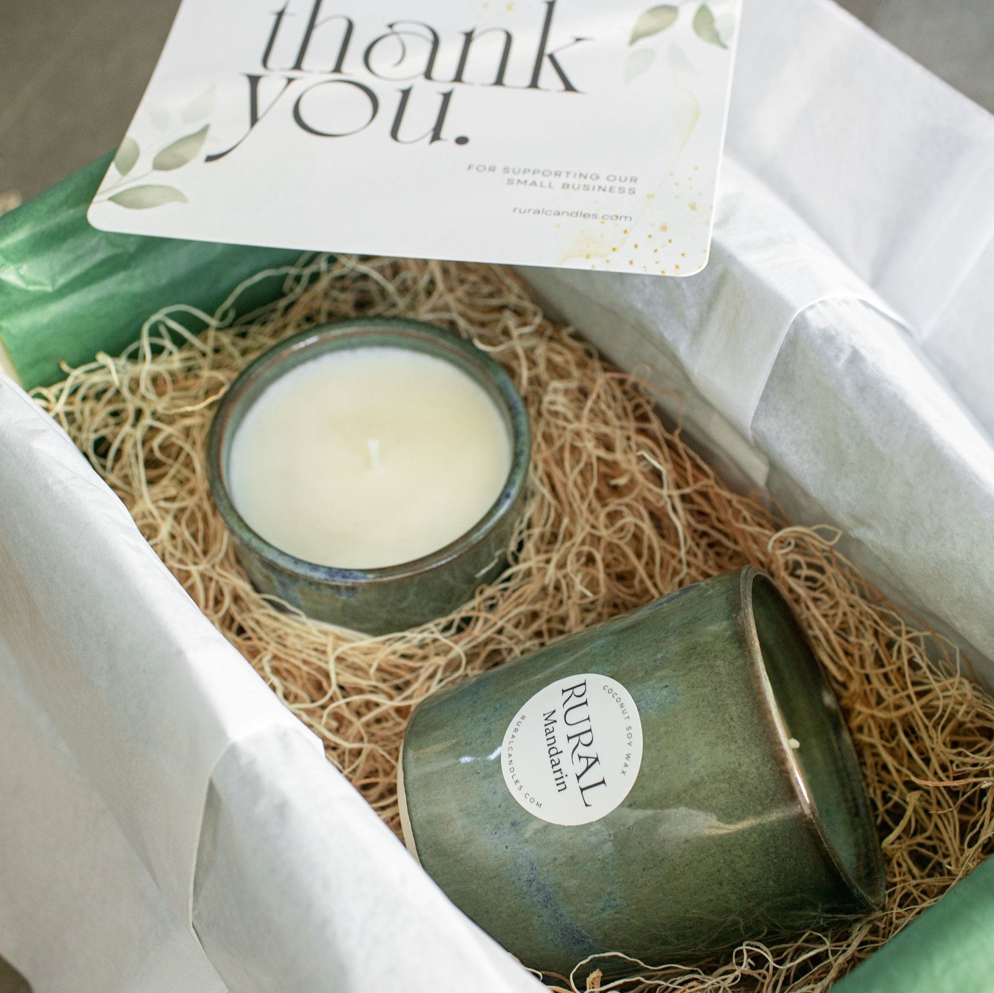 Photo of a pair of handcrafted pottery candles with a beautiful blue-green and brown glaze, nestled in a wooden nest within a gift box with a beautiful Thank You card alongside.