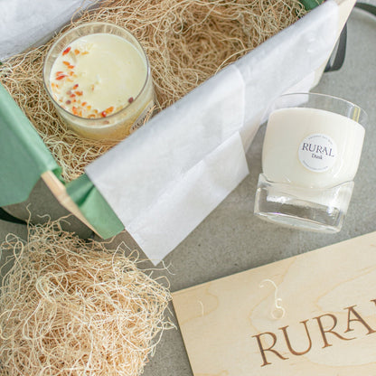 Photo of two very unique hand-blown, heavy bottom glass whiskey tumblers that have been handcrafted into a RURAL Candles scented candles with gemstones on top. They are presented in a wooden gift box, nestled in wood shavings. The mood is very clean, natural, elevated gifting.