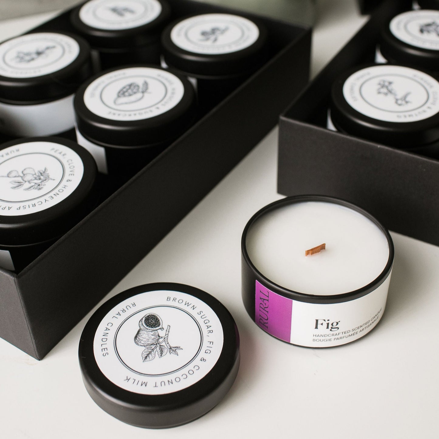 Candle with 'Fig' scent on a white surface, surrounded by black boxes and a plant.