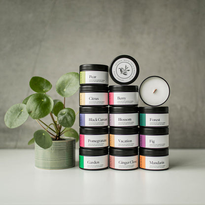 Stack of candles with colourfully labelled lids on a white surface next to a small plant, against a gray background.