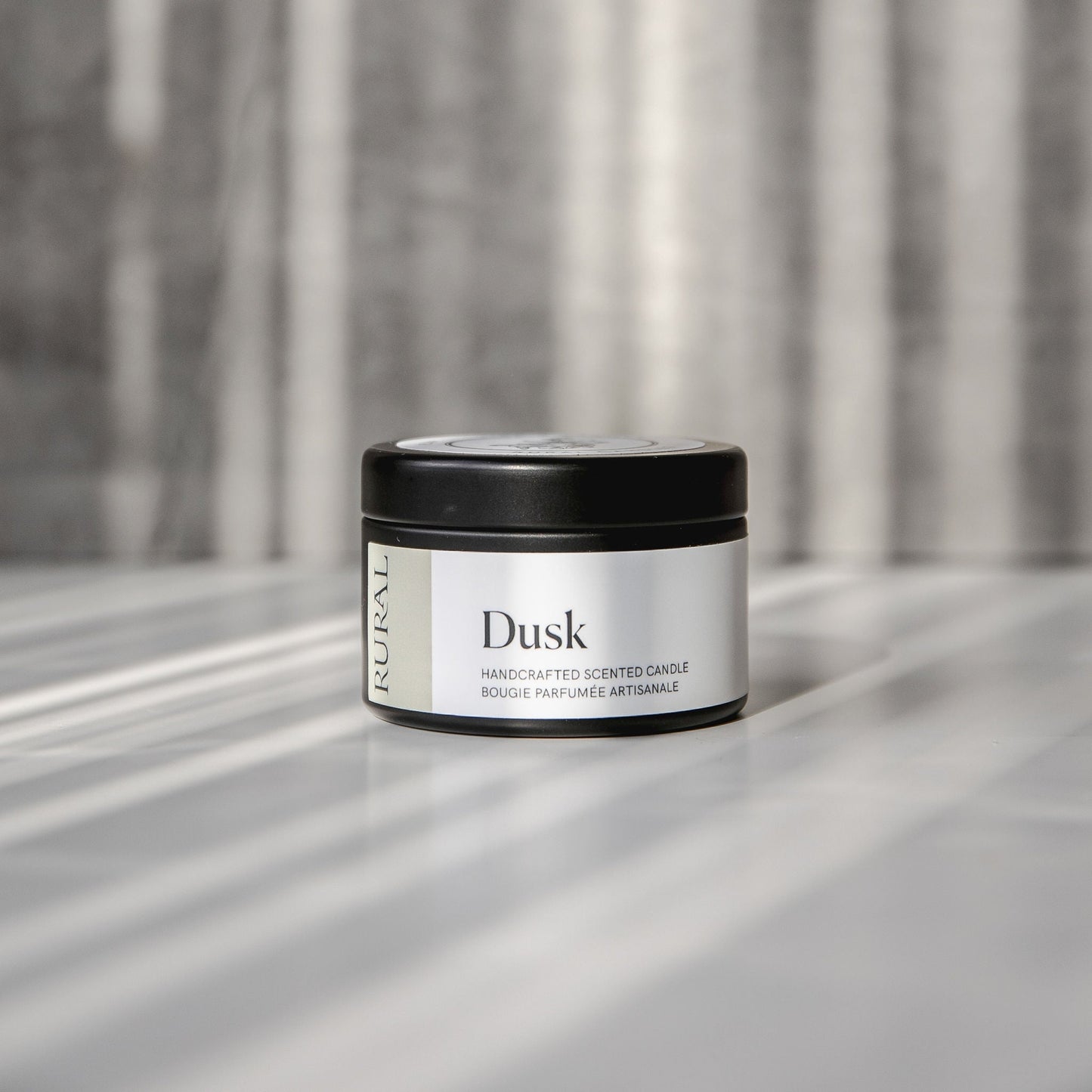 Dusk scented black candle tin with tan and white label on a reflective surface with a neutral background.