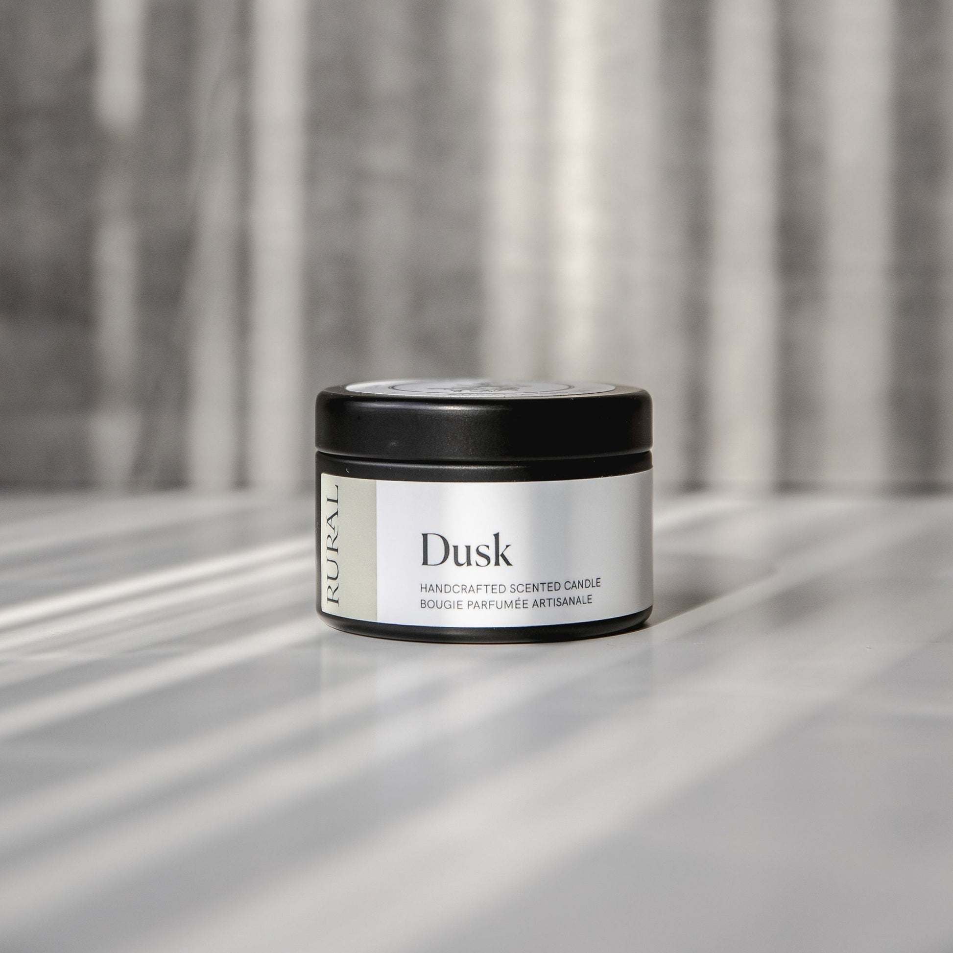 Dusk scented black candle tin with tan and white label on a reflective surface with a neutral background.
