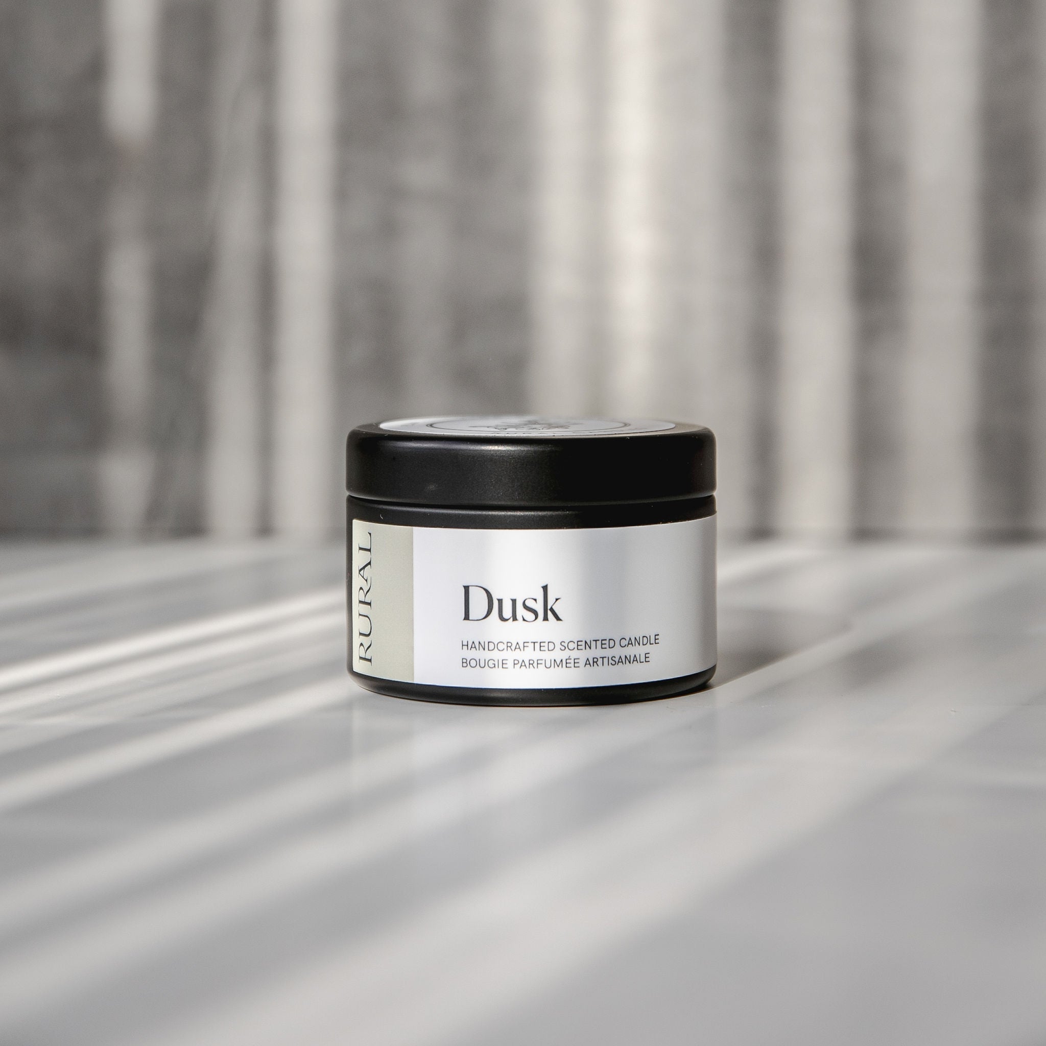 Dusk scented black candle tin with tan and white label on a reflective surface with a neutral background.