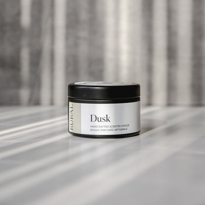 Dusk scented black candle tin with tan and white label on a reflective surface with a neutral background.