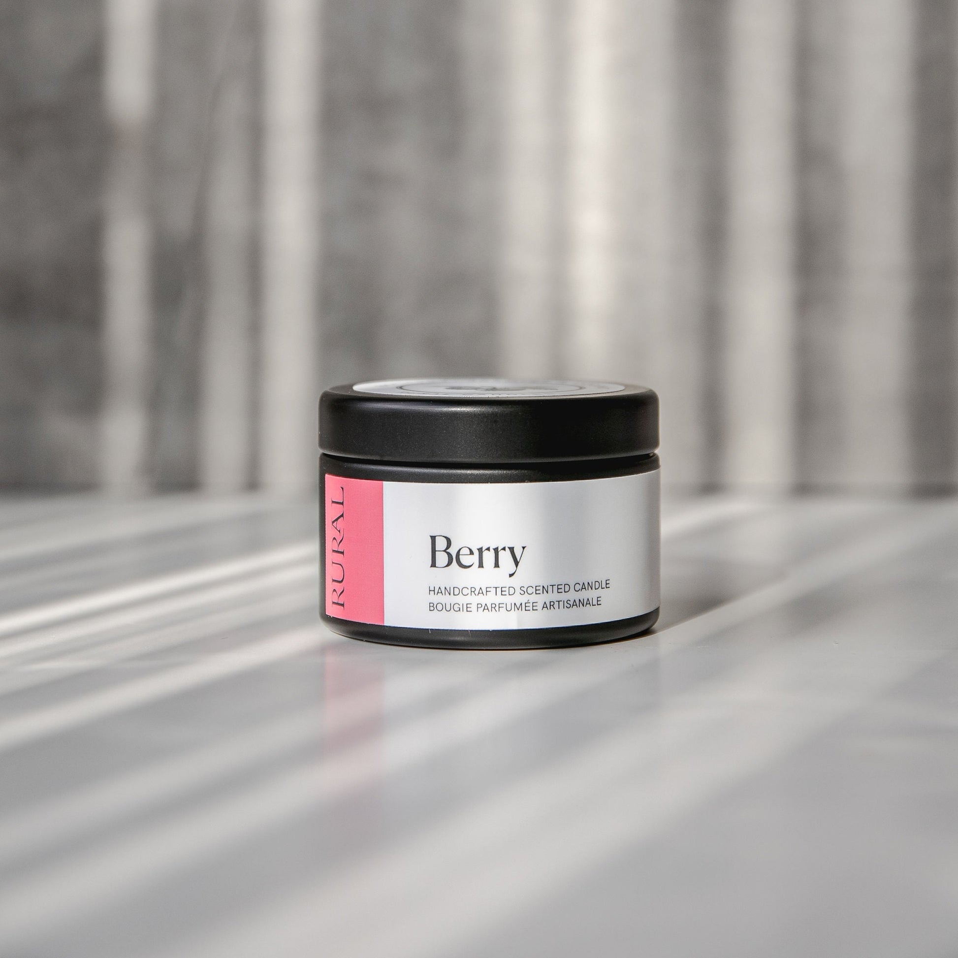 Berry scented black candle tin with pink and white label on a reflective surface with a neutral background.