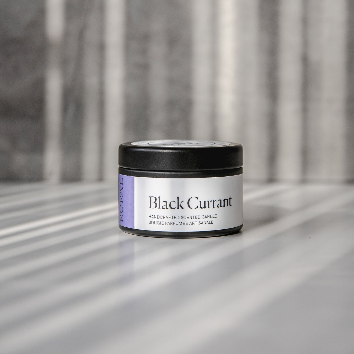 Black Currant scented black candle tin with purple and white label on a reflective surface with a neutral background.