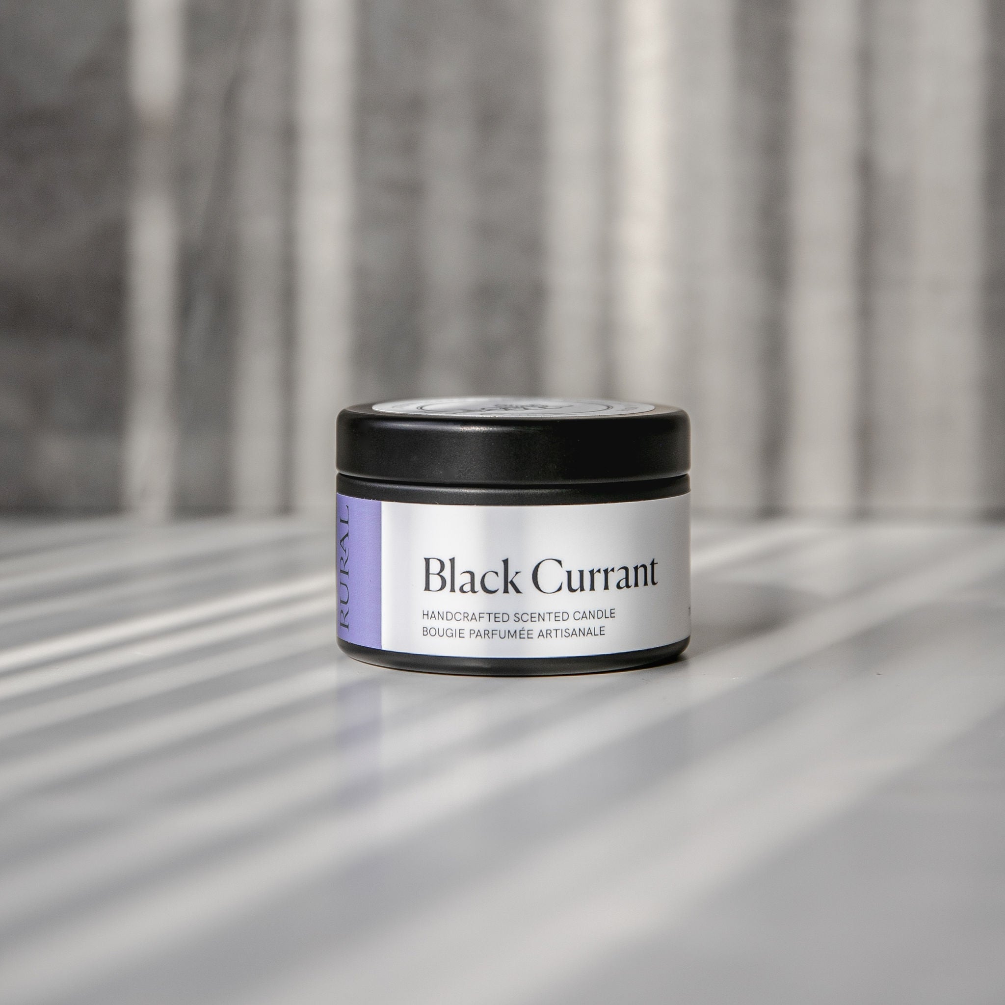 Black Currant scented black candle tin with purple and white label on a reflective surface with a neutral background.