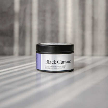 Black Currant scented black candle tin with purple and white label on a reflective surface with a neutral background.