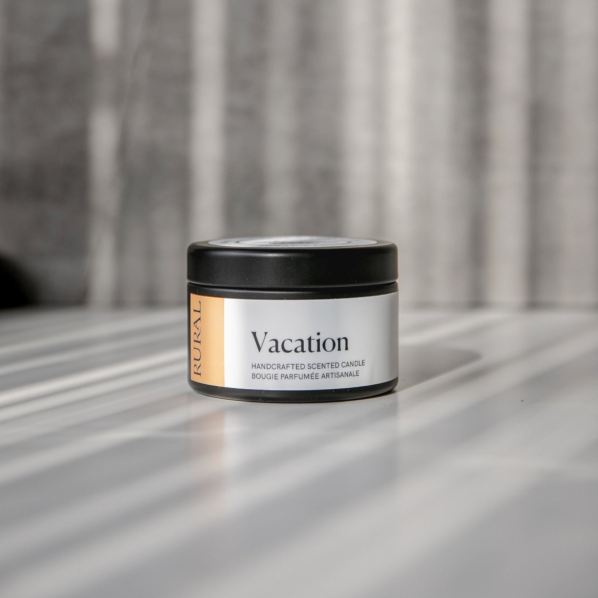 Vacation scented black candle tin with orange and white label on a reflective surface with a neutral background.