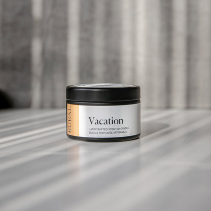 Vacation scented black candle tin with orange and white label on a reflective surface with a neutral background.