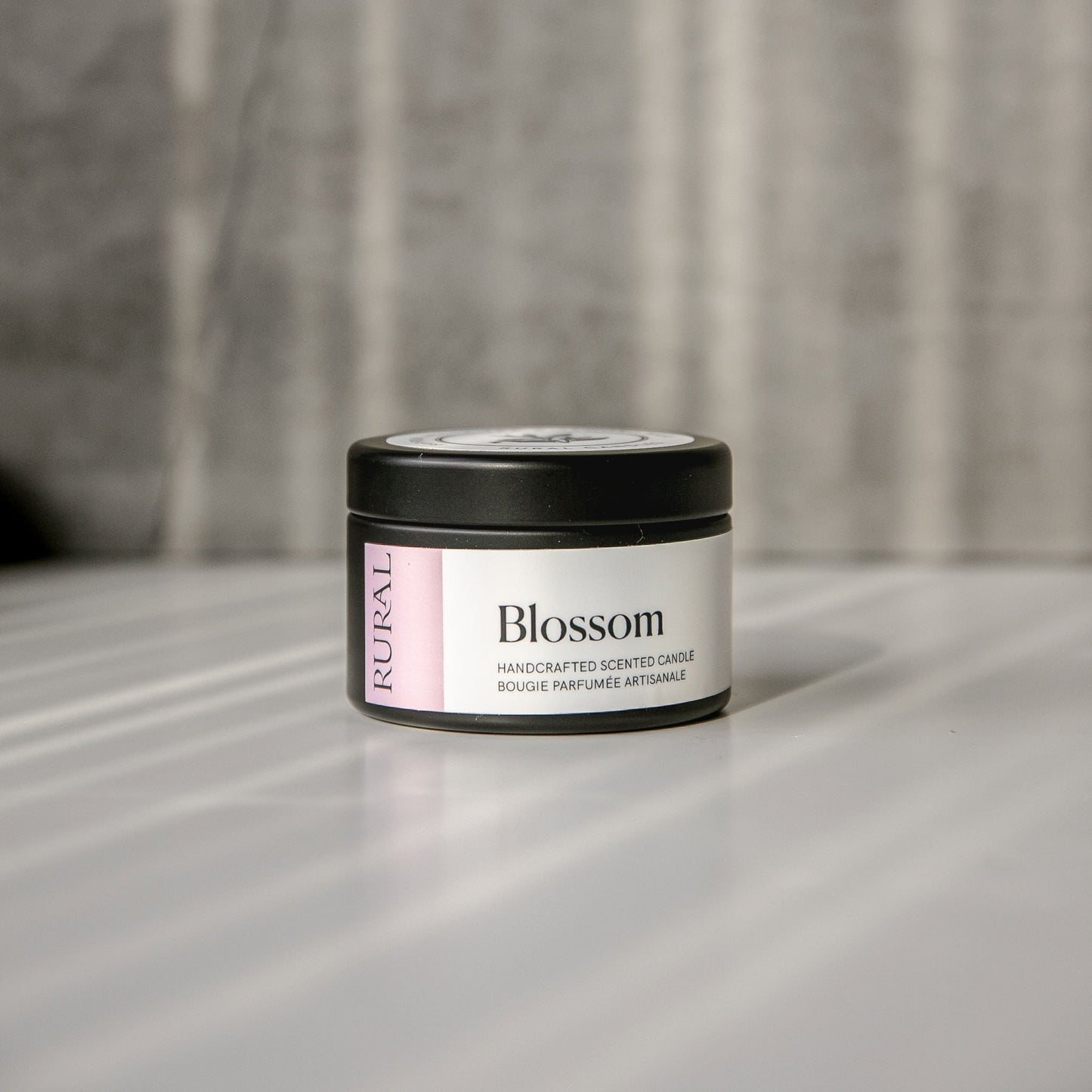 Blossom scented black candle tin with lilac and white label on a reflective surface with a neutral background.
