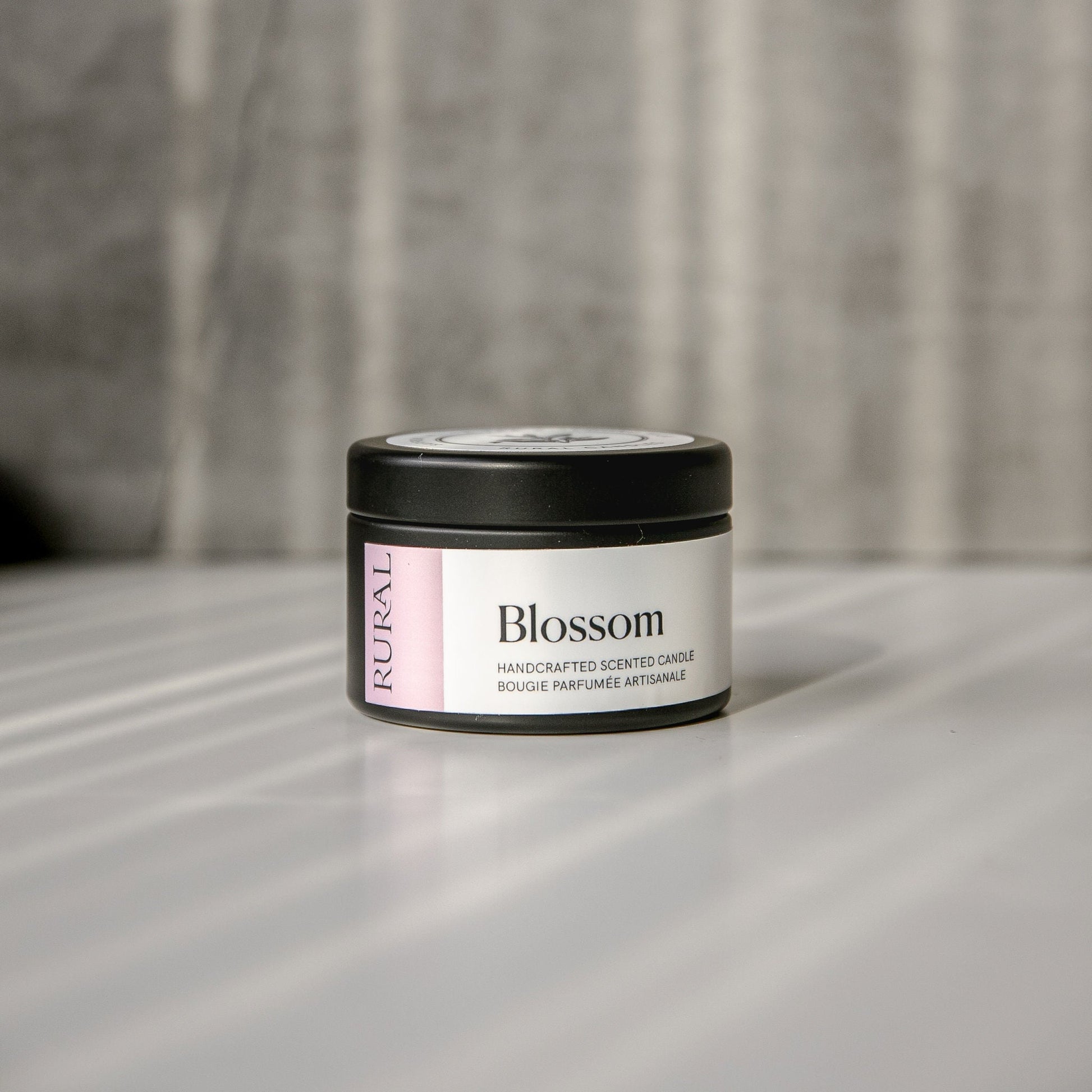 Blossom scented black candle tin with lilac and white label on a reflective surface with a neutral background.