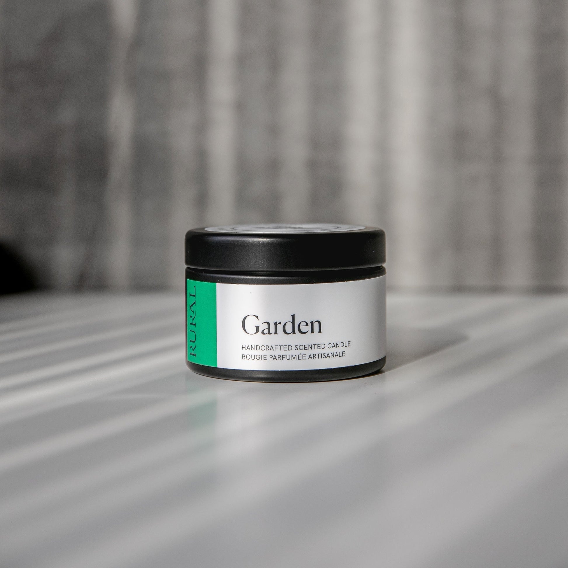 Garden scented black candle tin with green and white label on a reflective surface with a neutral background.