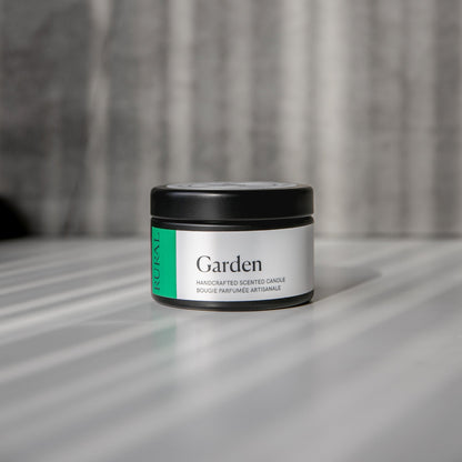 Garden scented black candle tin with green and white label on a reflective surface with a neutral background.