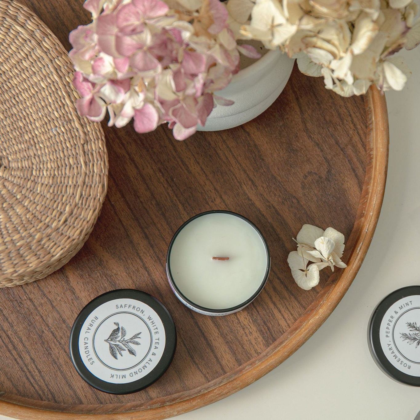 Candles with decorative labels on a wooden tray with flowers and a box of candles.