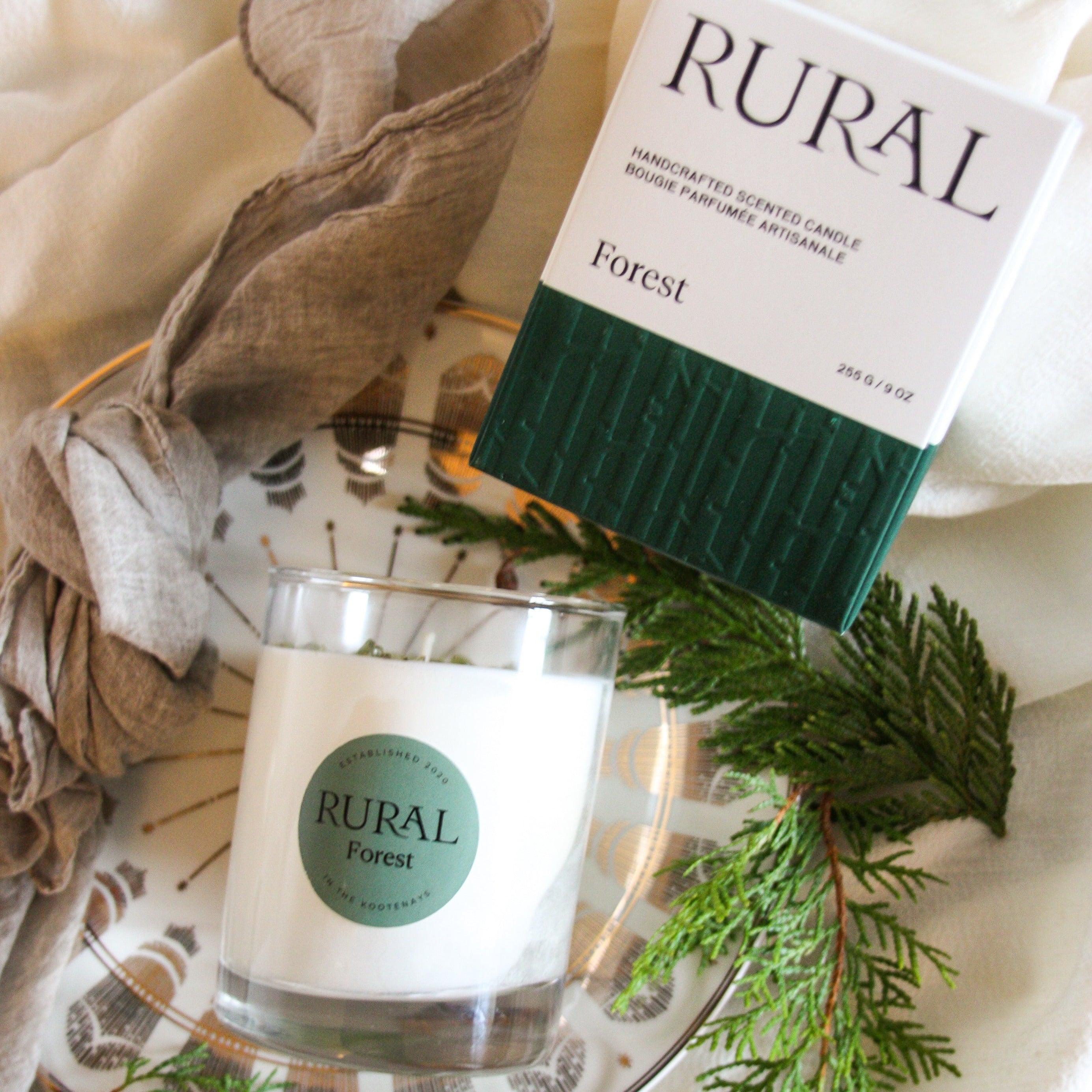 Close-up photo of a white candle in clear glass with a round green label indicating RURAL Candles Forest Scent beside its matching green and white elegantly embossed candle gift box. The candle is arranged on a gold and white ceramic plate, with earthy-toned linens and cedar branches alongside. 