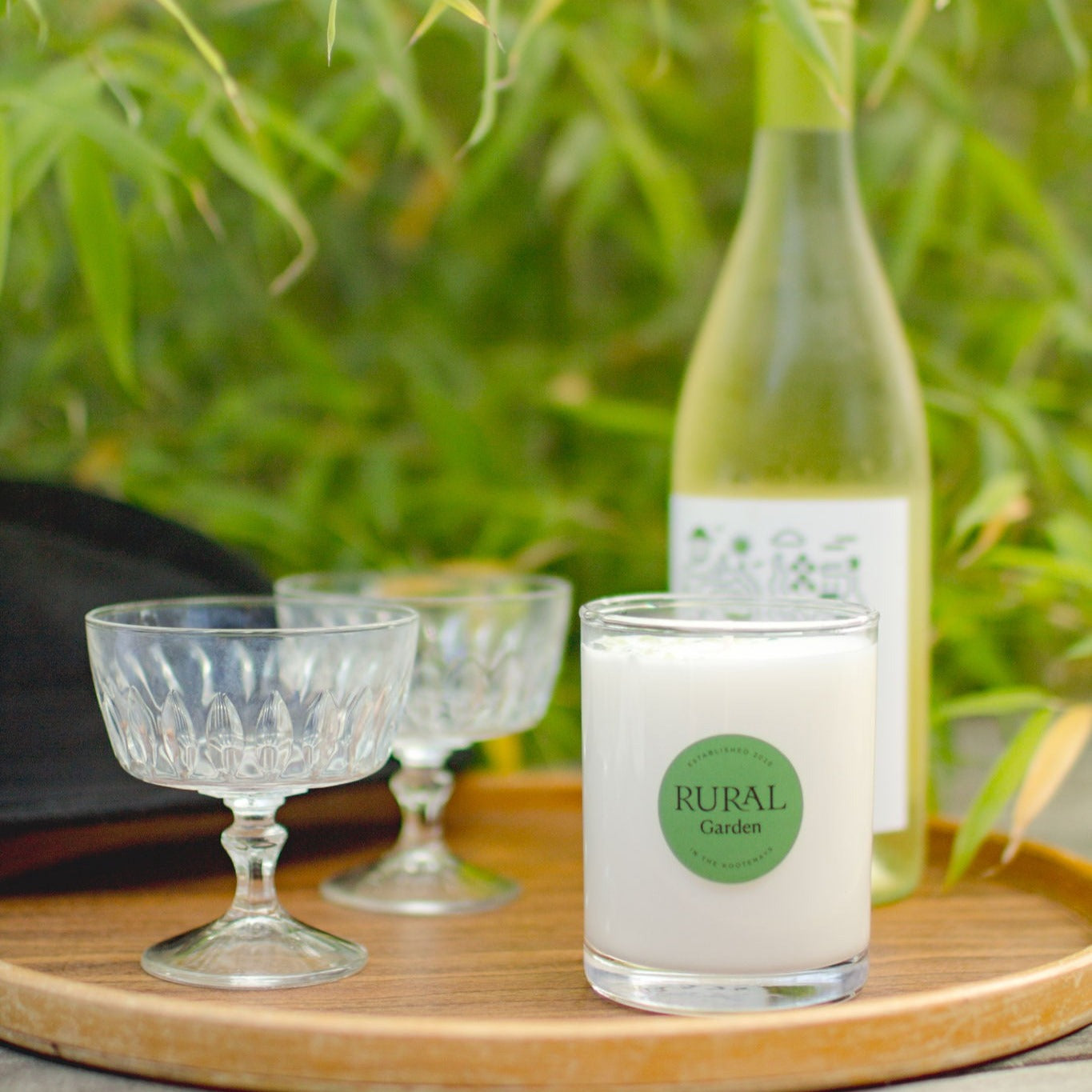 Photo of a coconut soy candle in a clear white glass with a round green label indicating RURAL Candles Garden Scent. The candle is sitting on a wooden tray with a bottle of white wine and glasses. The scene is a summer garden party in a lush, green garden. 