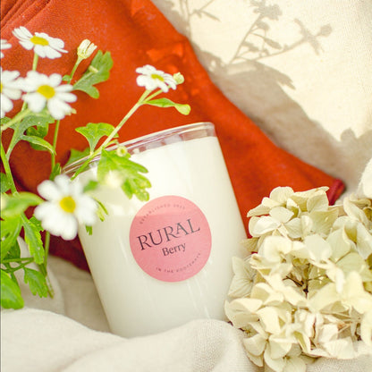 Photo of a white candle in a clear glass container with a pink round RURAL Candles label on it, indicating its Berry scent. The candle is nestled in white and red linen fabric with white flowers and sunshine surrounding it.