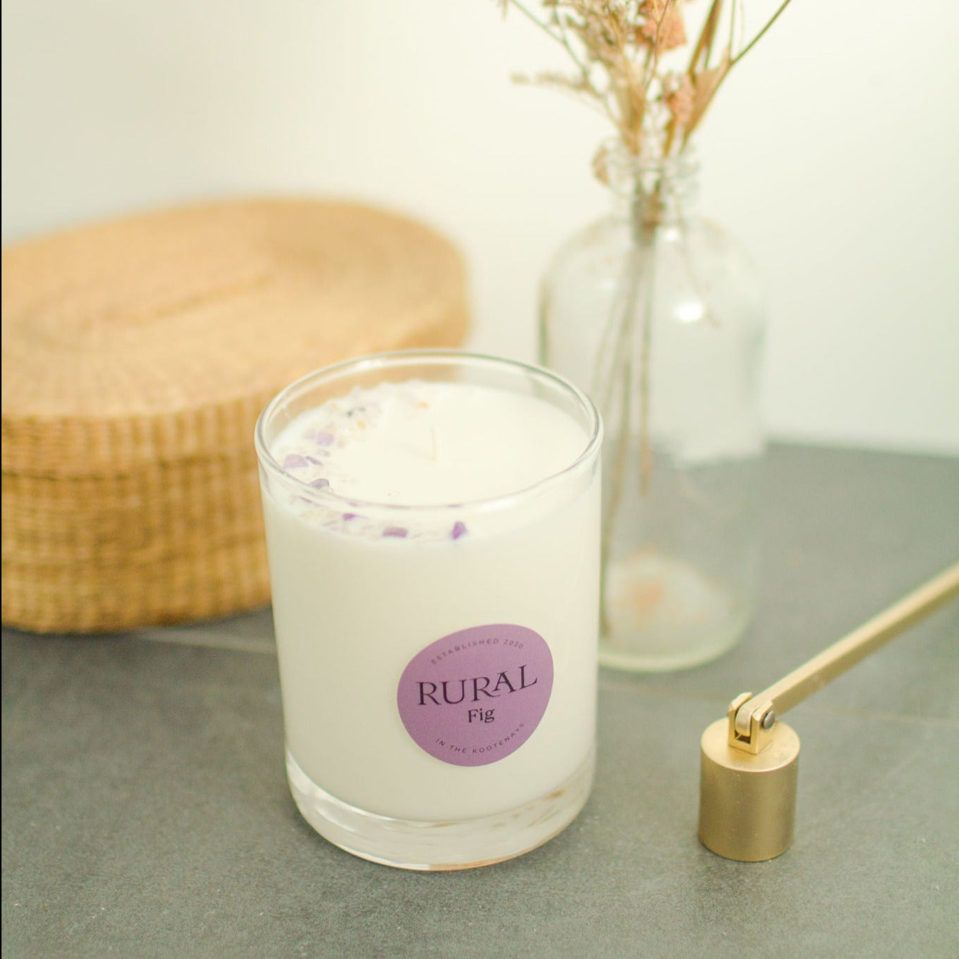 Photo of a white candle in a clear glass with a round purple label indicating RURAL Candles Fig scent. We can see the top of the candle is decorated with amethyst gemstones arranged in a crescent moon shape. The candle is slate counter, with a gold candle snuffer beside it and a wicker basket and flowers in the background.